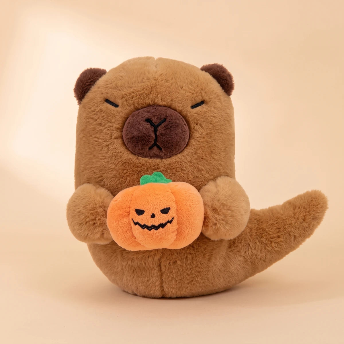 Front view of the Halloween ghost capybara plush holding its orange pumpkin on a peach studio backdrop