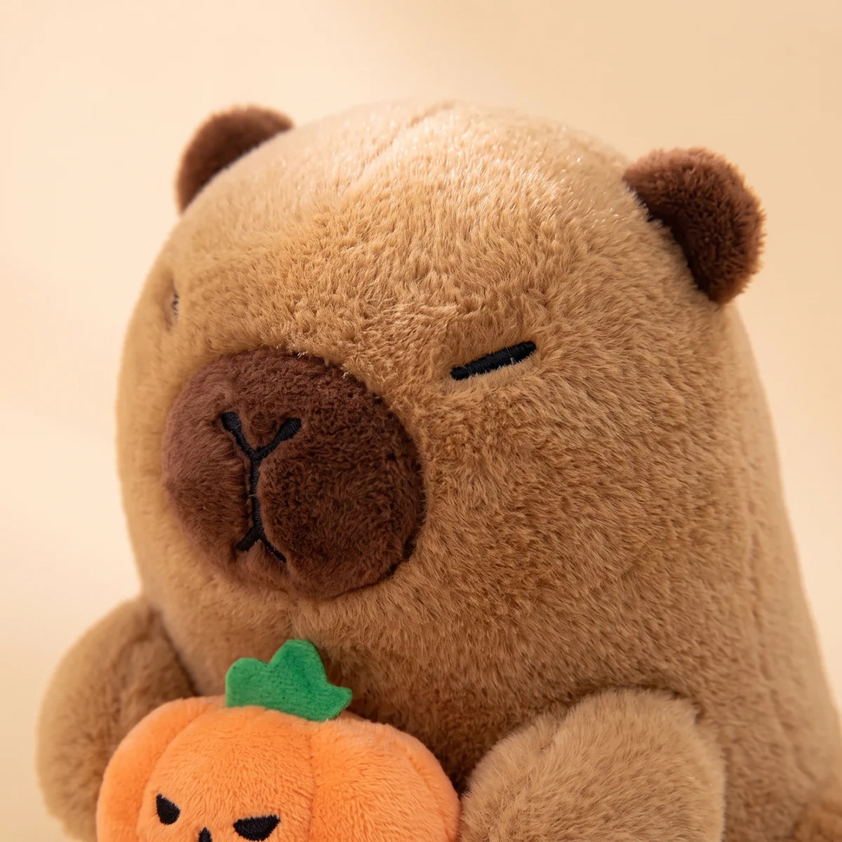 Close-up of the ghost capybara plush face with stitched closed eyes above its pumpkin's green leaf