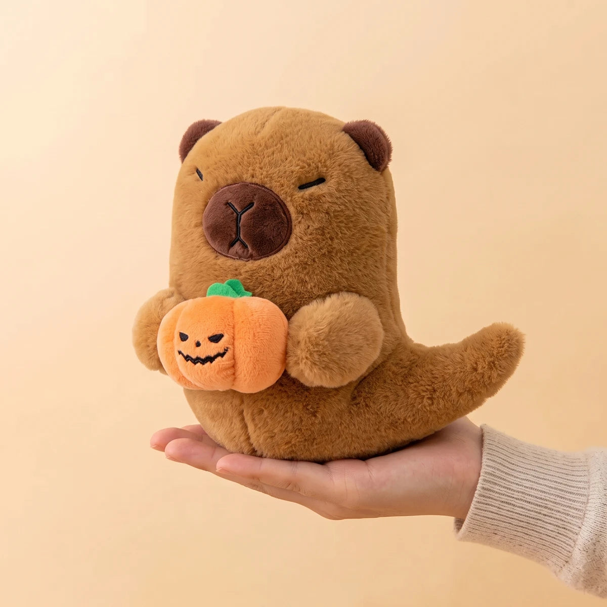 Ghost capybara plush with pumpkin resting on an open hand to show its 7 in size