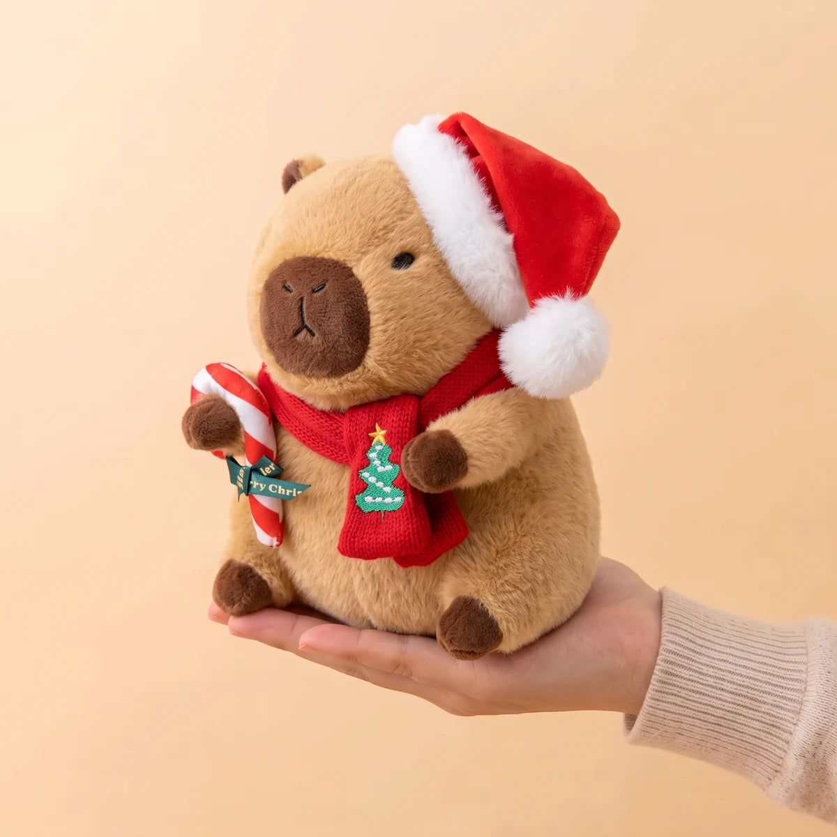 9 in Christmas capybara plush with a Santa hat holding a candy cane sitting on an open hand