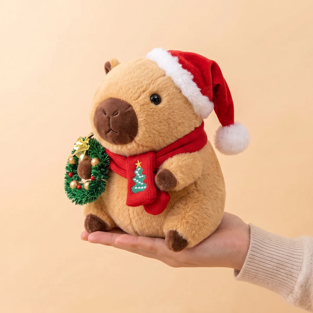 9 in Christmas capybara plush with a Santa hat holding a green wreath sitting on an open hand