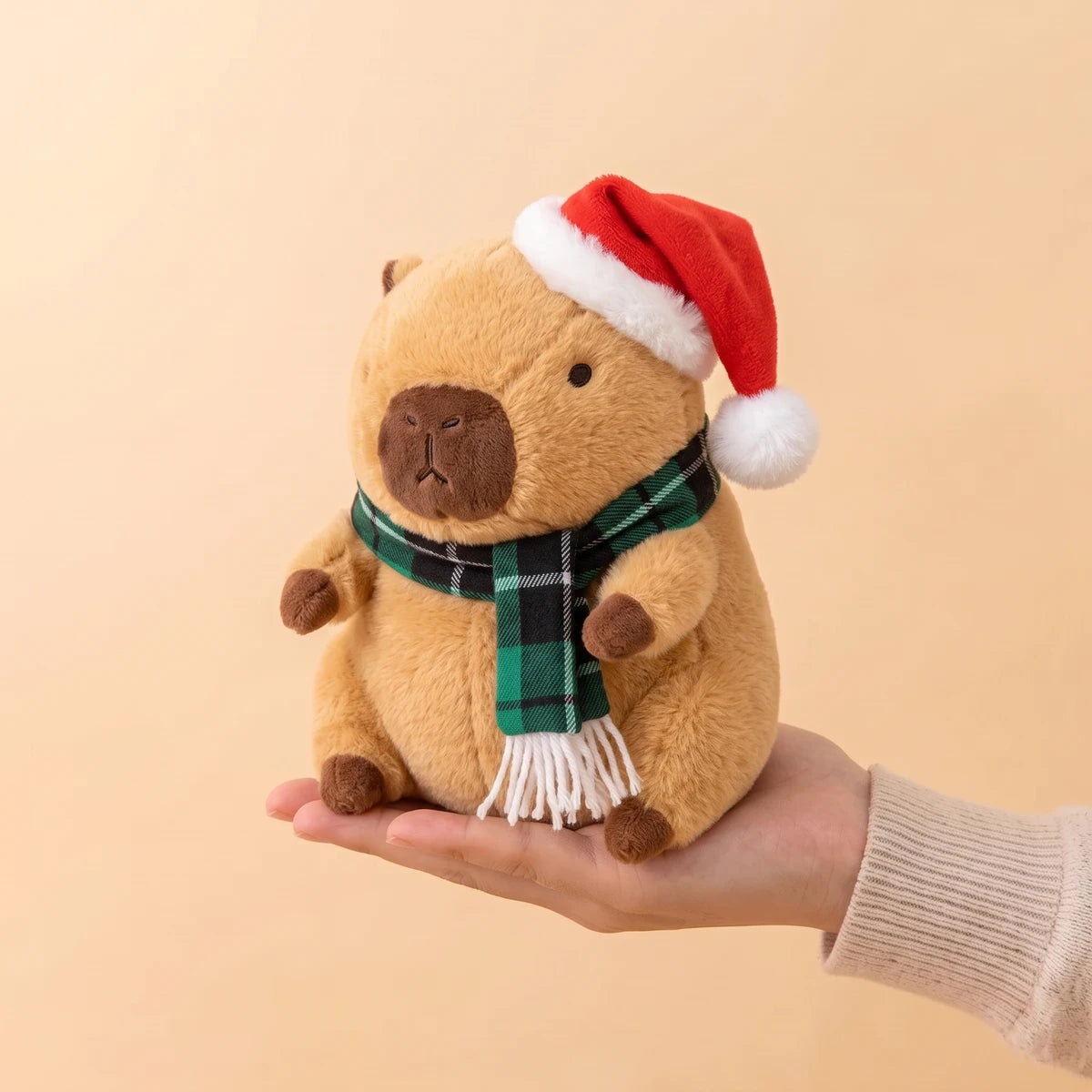 9 in Christmas capybara plush with a Santa hat and a green plaid scarf sitting on an open hand
