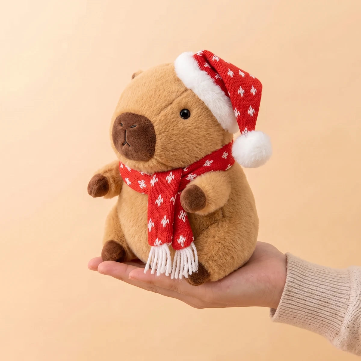 9 in Christmas capybara plush with a red knit hat and scarf dotted with snowflakes sitting on an open hand