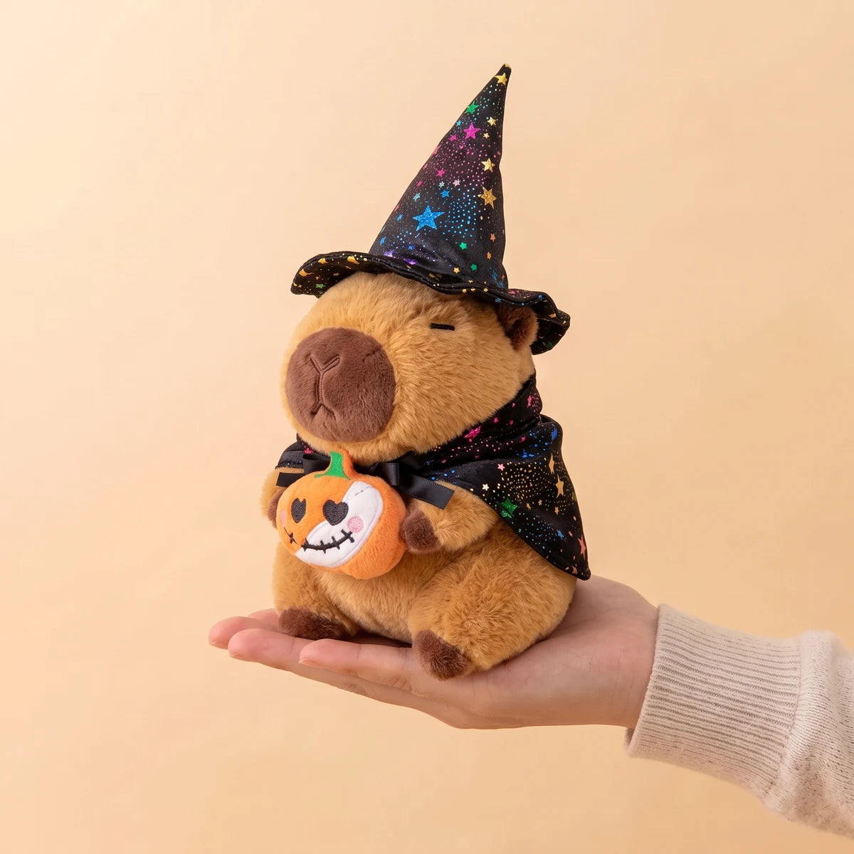 Pumpkin witch capybara plush holding a small pumpkin, sitting on an open hand