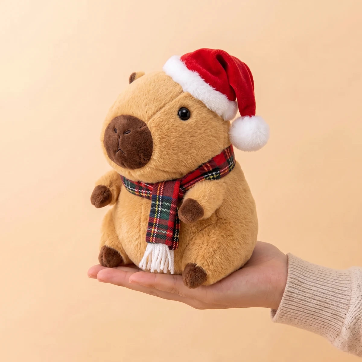9 in Christmas capybara plush with a Santa hat and a red tartan scarf sitting on an open hand