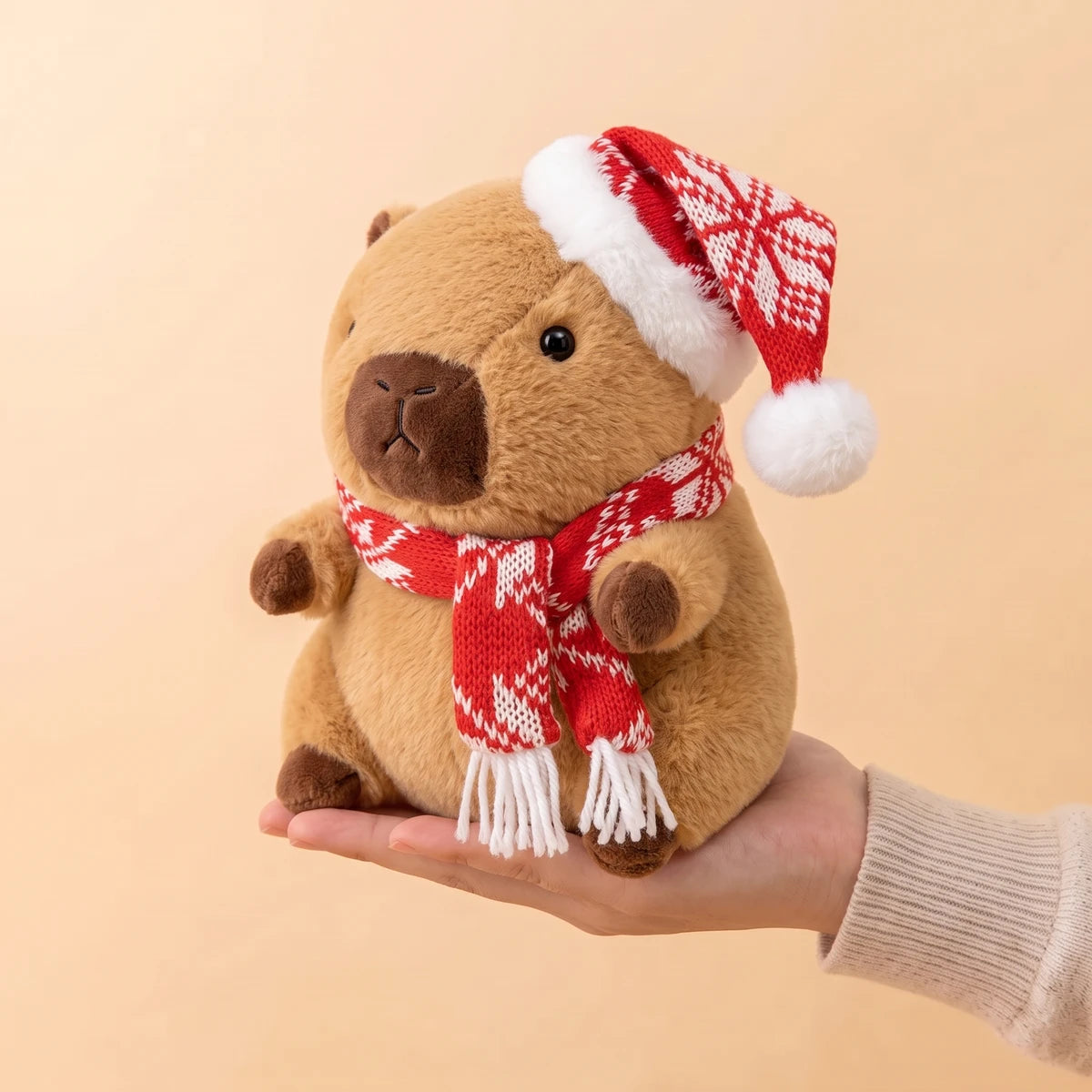 9 in Christmas capybara plush with a red snowflake knit hat and scarf sitting on an open hand