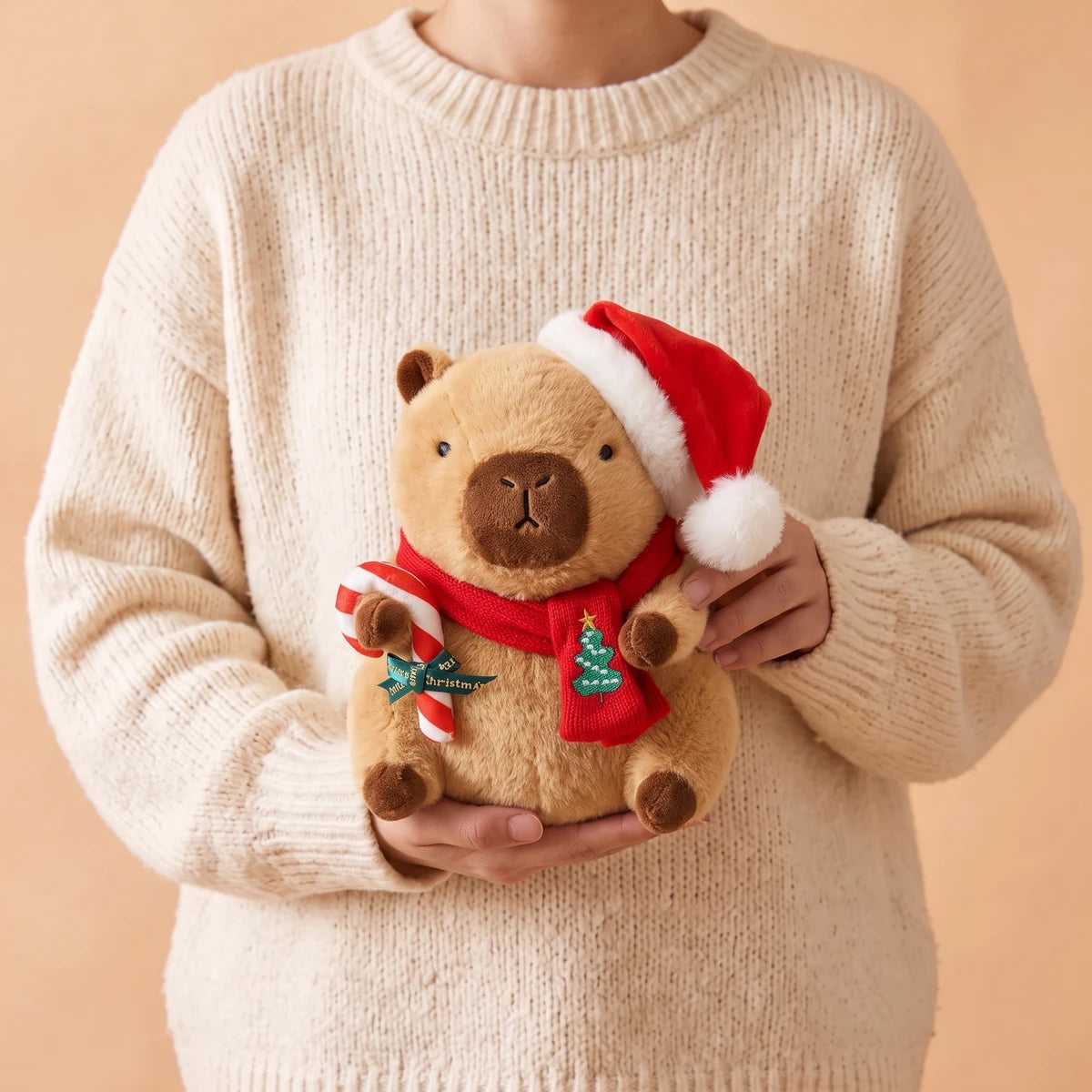 Person in a cream sweater holding the 9 in Christmas capybara plush with a Santa hat holding a candy cane