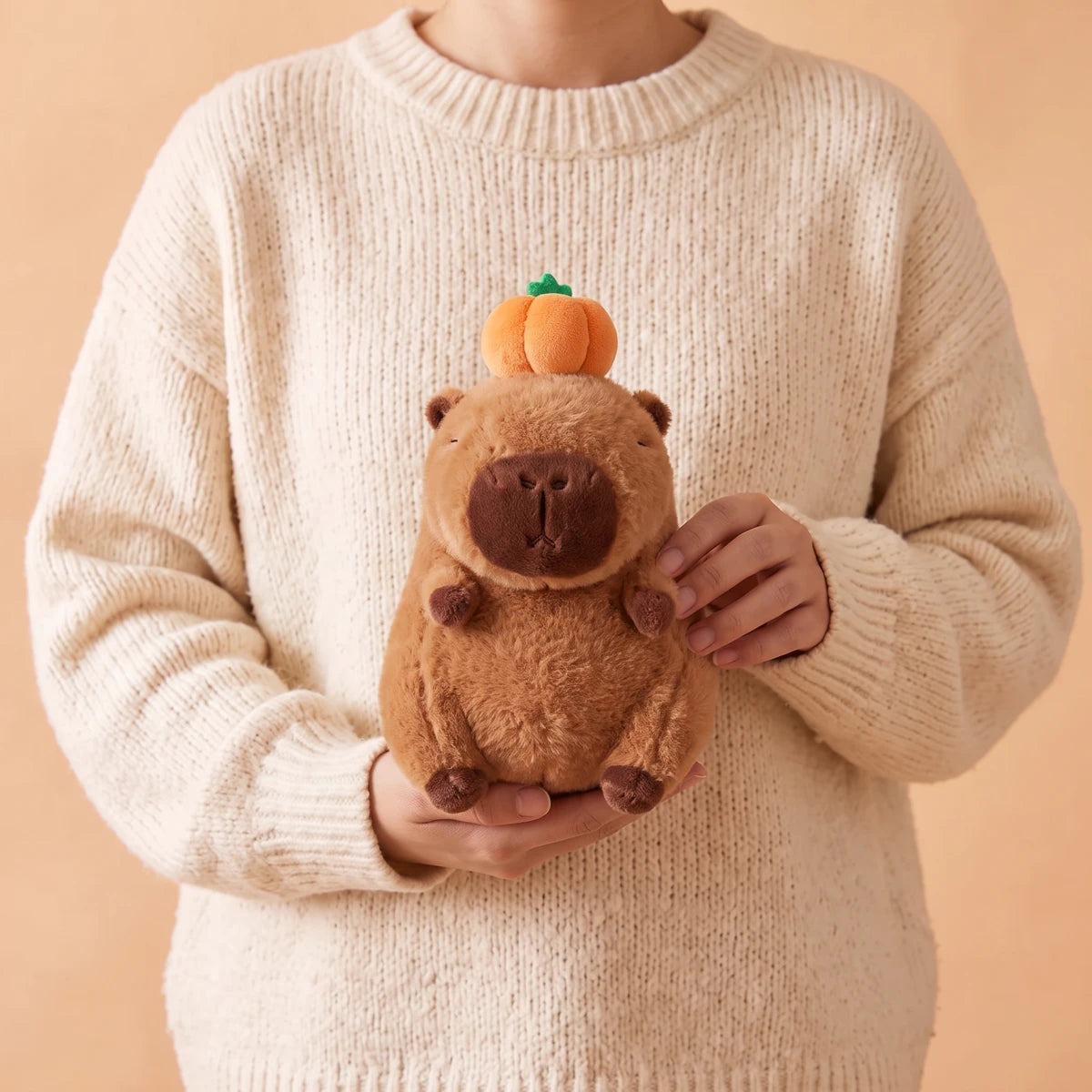 Person in a cream sweater holding the capybara with pumpkin on head plush