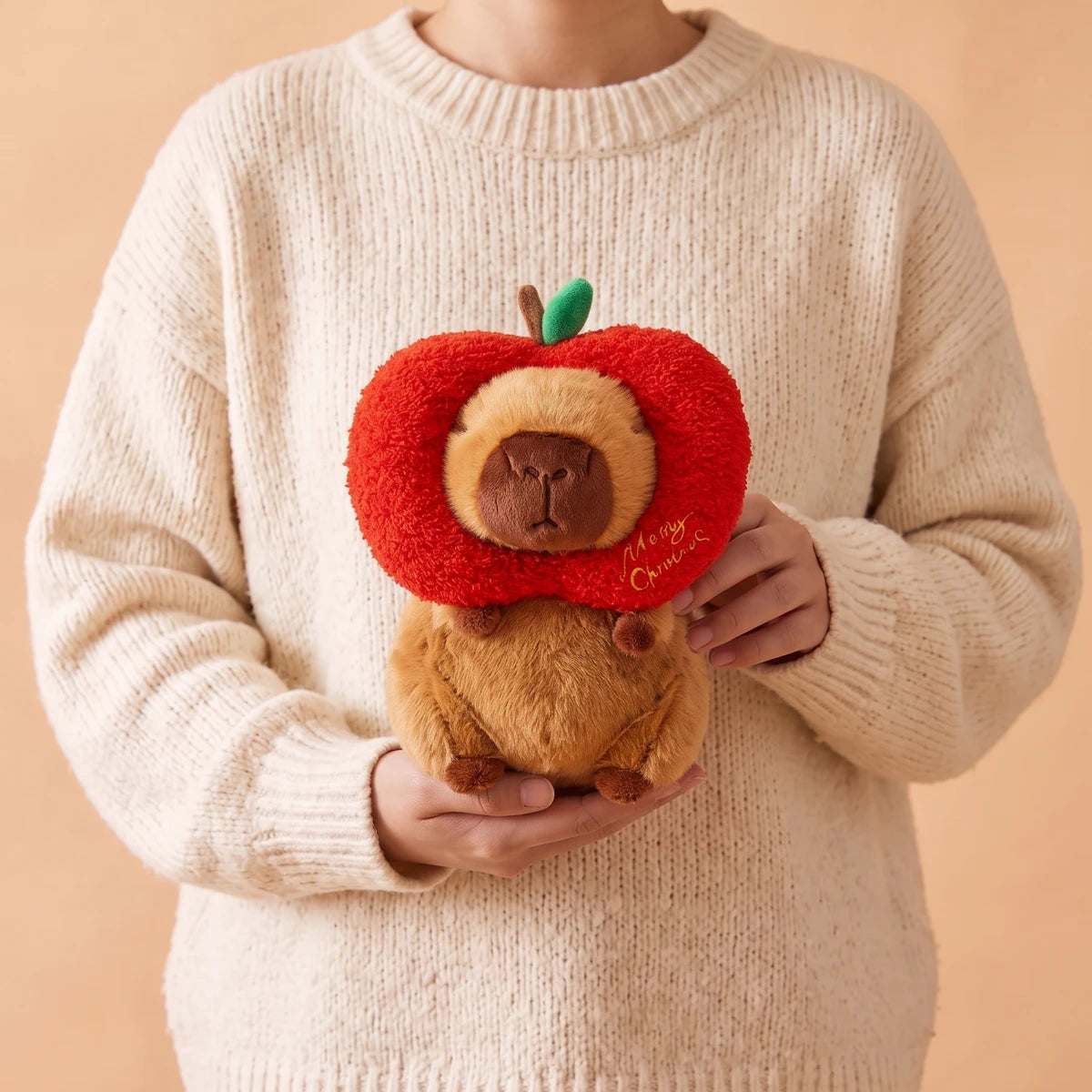 Person in a cream sweater holding the 8 in Christmas apple capybara plush