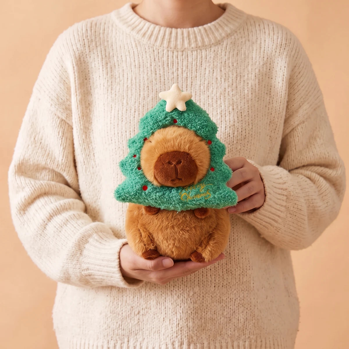 Person in a cream sweater holding the 8 in Christmas tree capybara plush