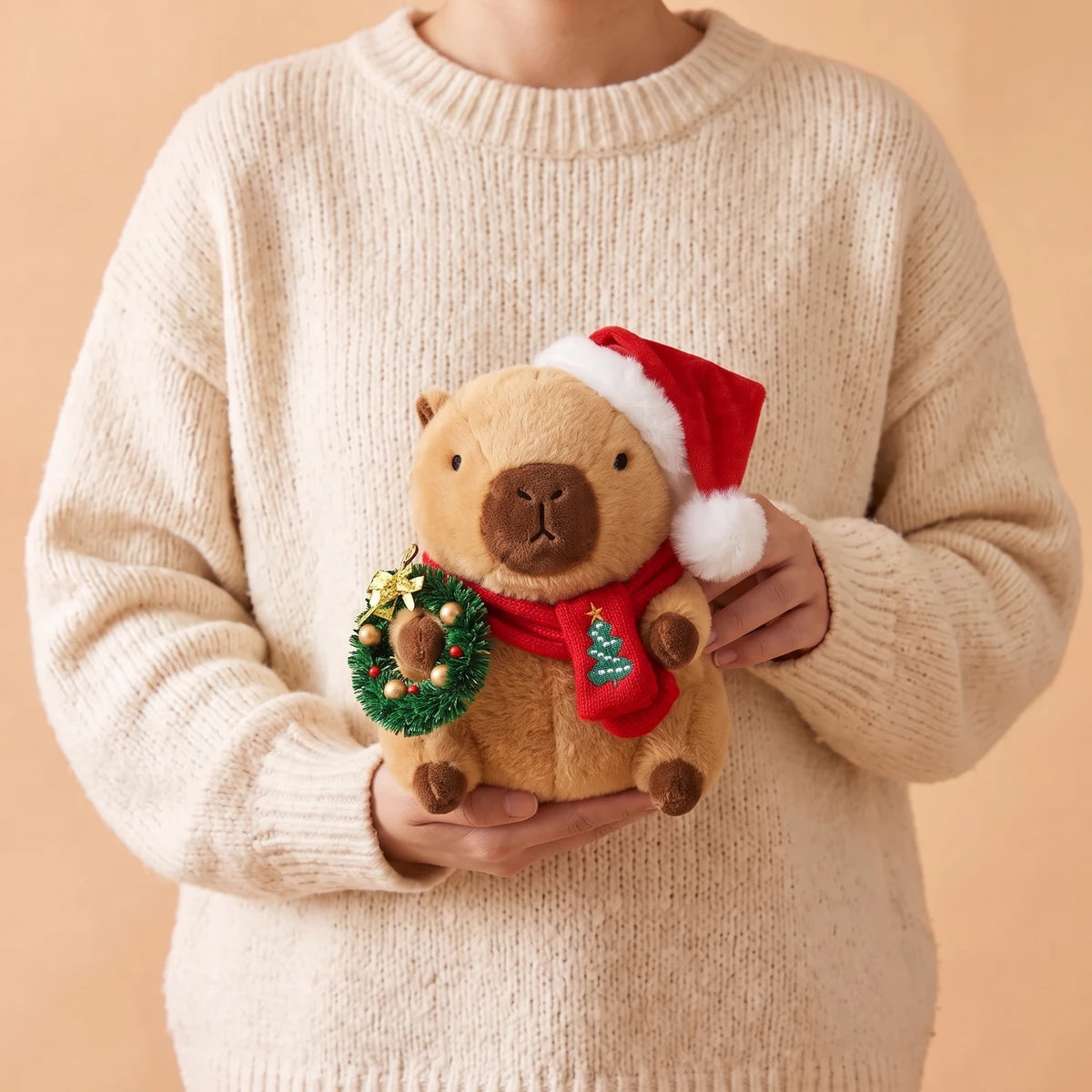 Person in a cream sweater holding the 9 in Christmas capybara plush with a Santa hat holding a green wreath