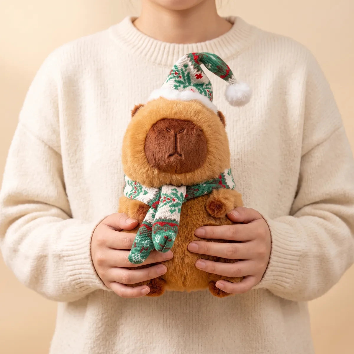 Person in a cream sweater holding the 10 in Christmas capybara plush with a green and white knit hat and scarf