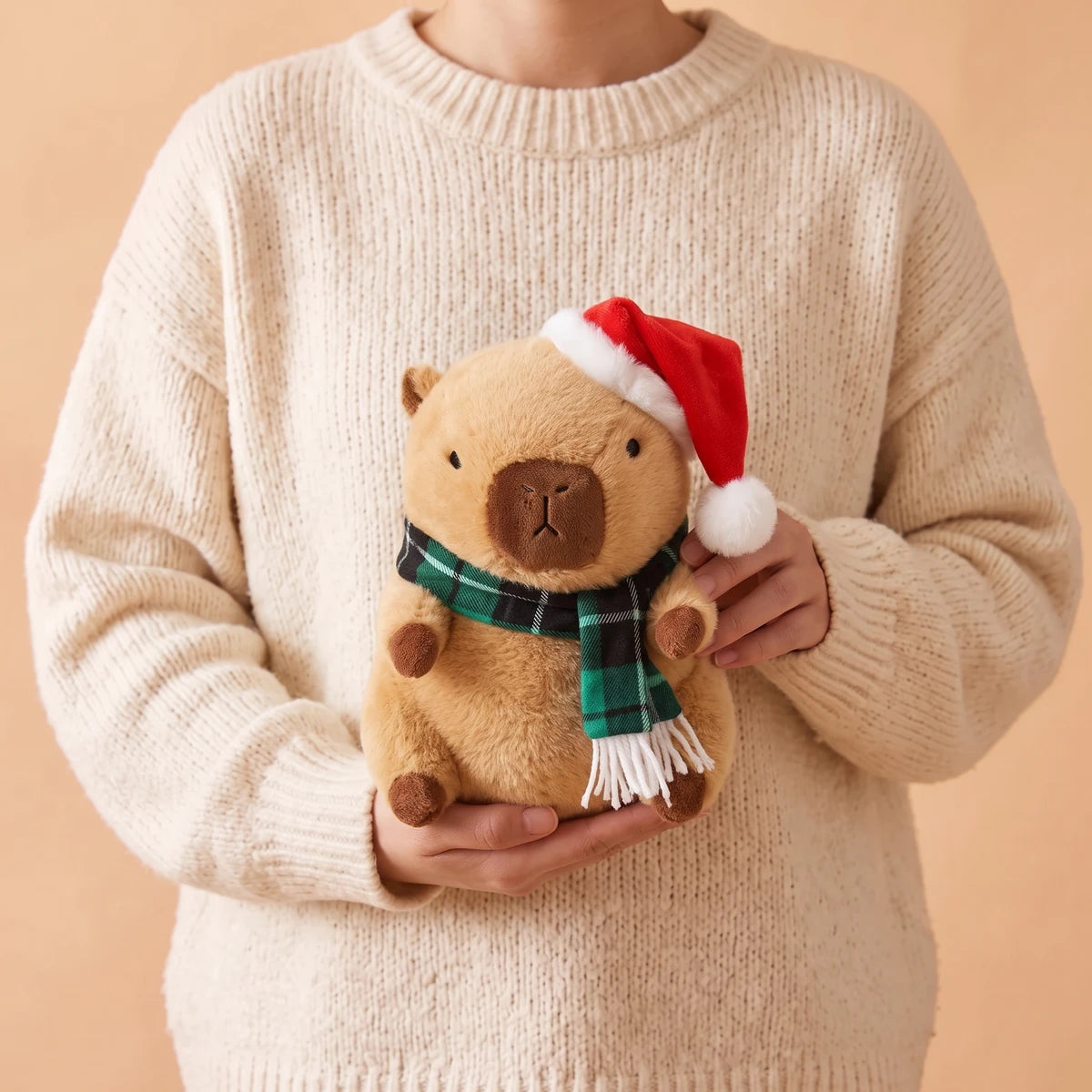 Person in a cream sweater holding the 9 in Christmas capybara plush with a Santa hat and a green plaid scarf