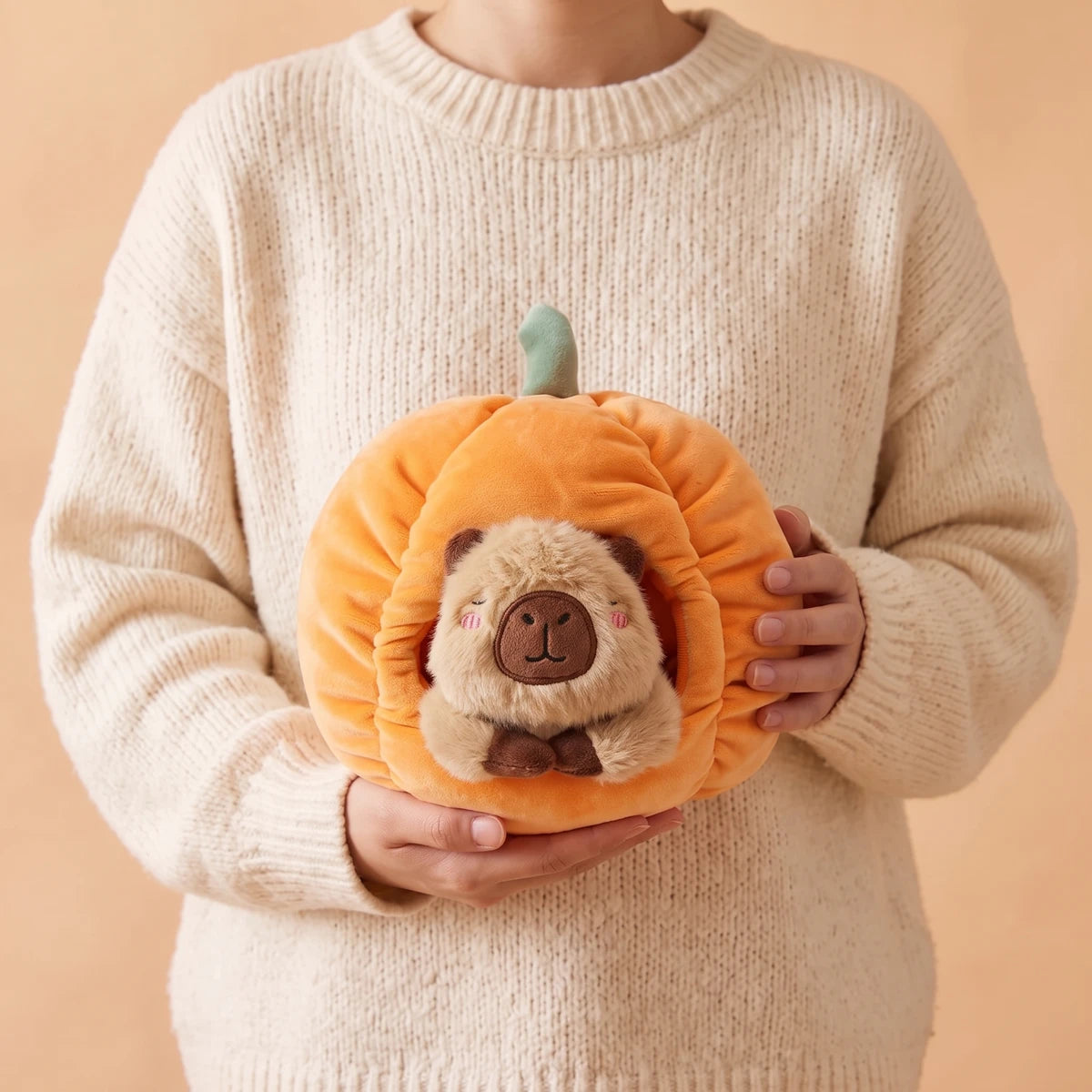 Person in a cream sweater holding the pumpkin house capybara plush set