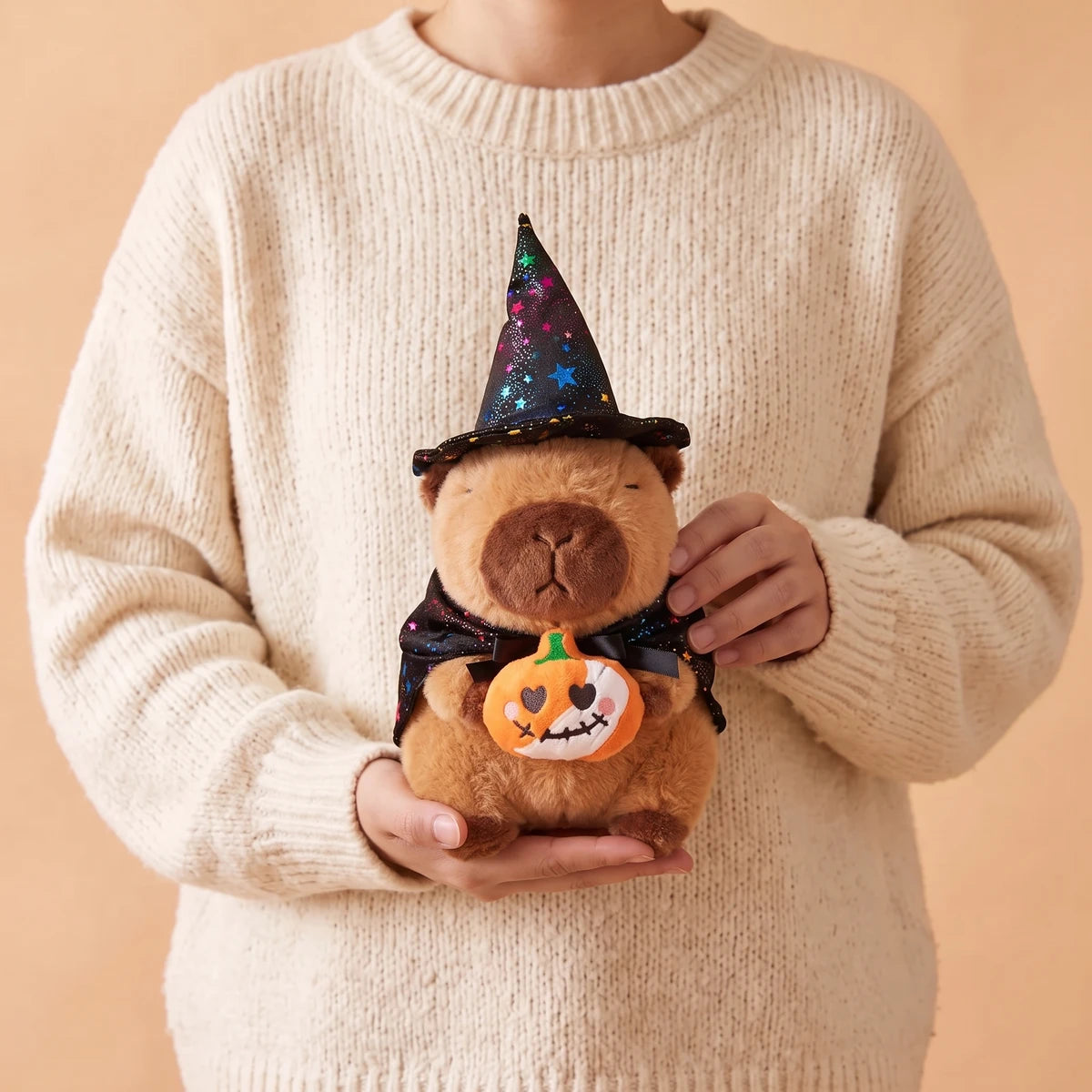 Person in a cream sweater holding the pumpkin witch capybara plush