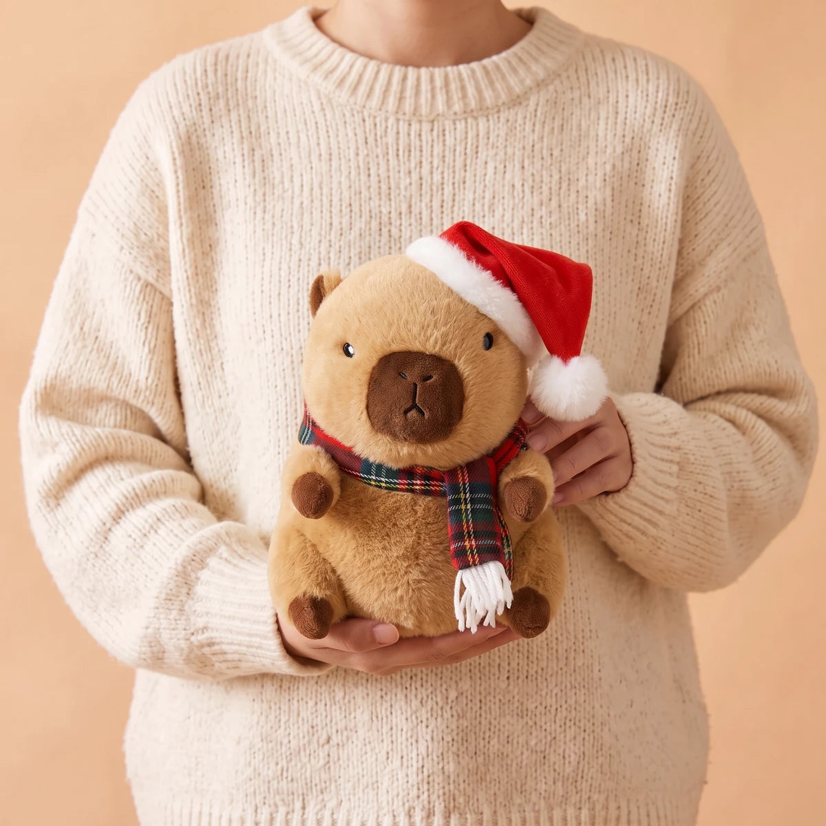 Person in a cream sweater holding the 9 in Christmas capybara plush with a Santa hat and a red tartan scarf