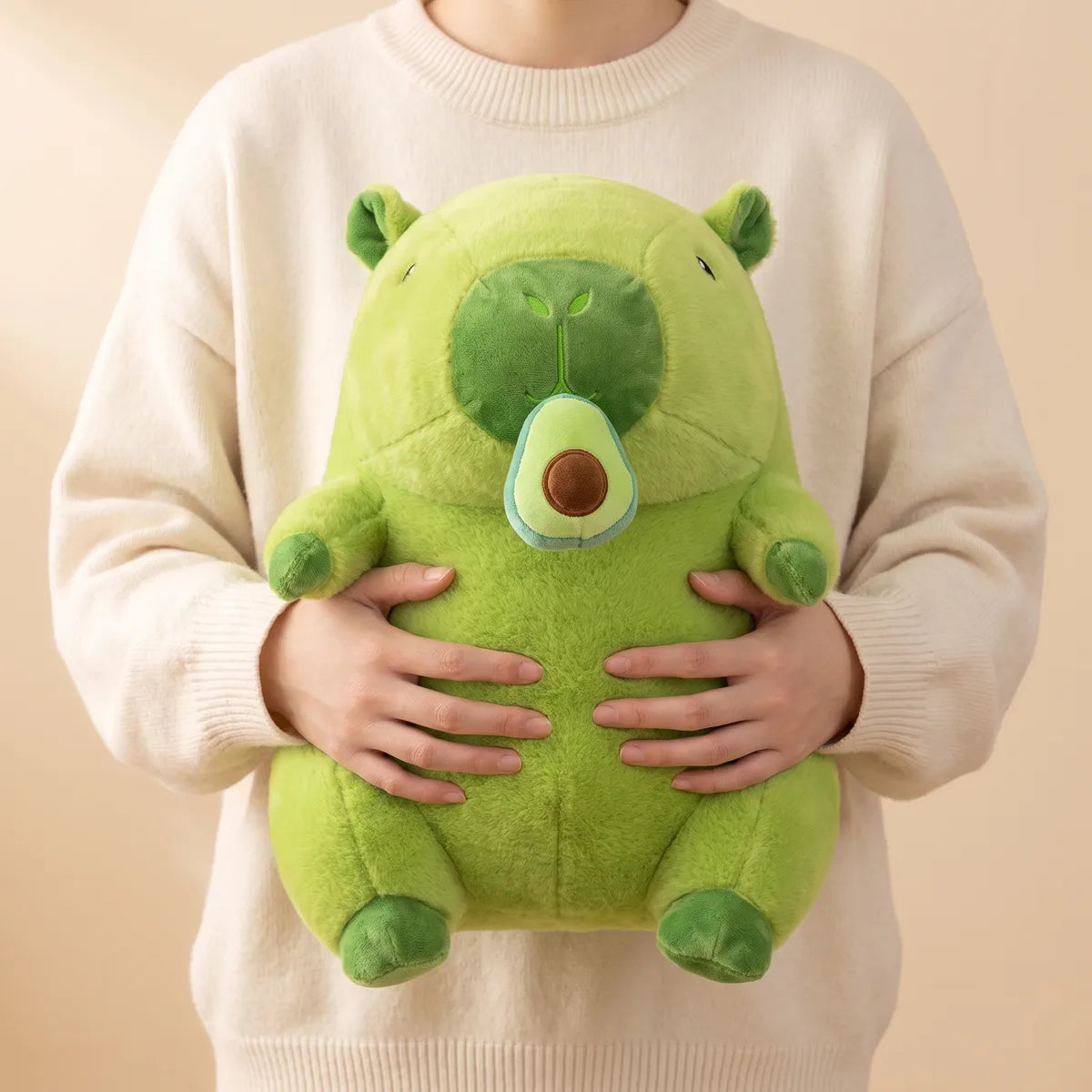 Person in a cream sweater holding the green capybara plush flat against their chest with both hands