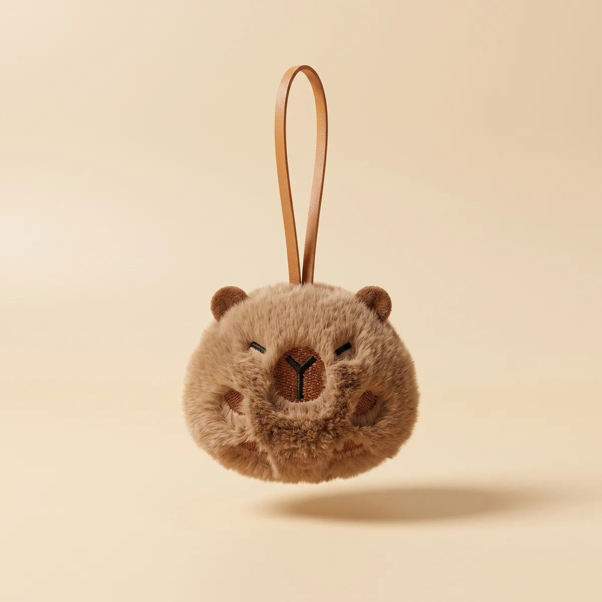 Ball capybara keychain hanging from its tan strap seen from the front