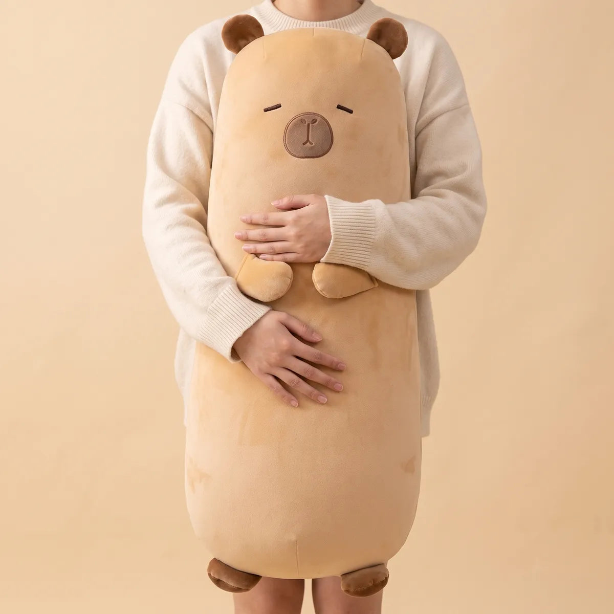 Big capybara stuffed animal plush held upright against an adult, the sand colored body reaching from mid thigh to shoulder