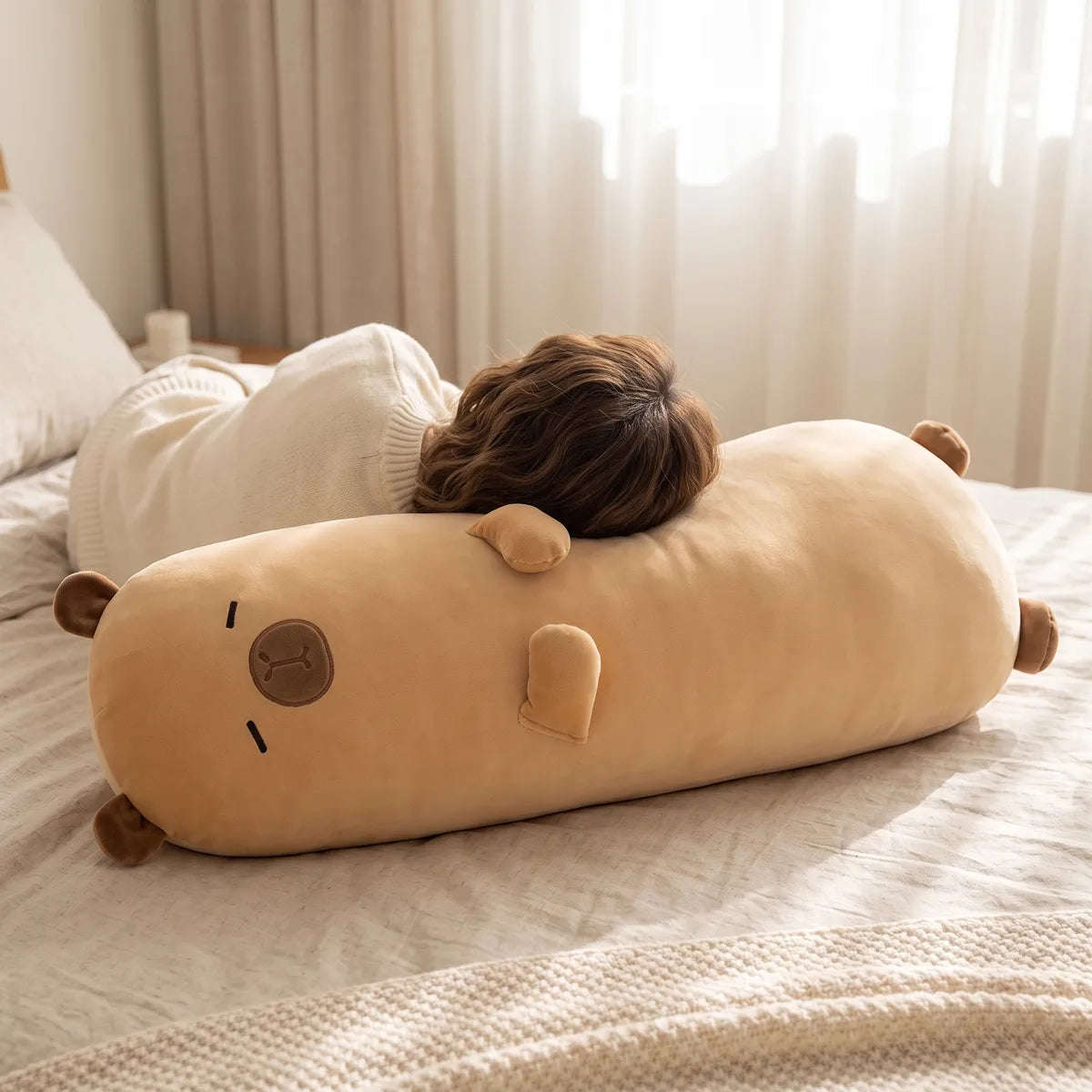 Big capybara stuffed animal plush laid on its side on a bed while a person rests their head against it.