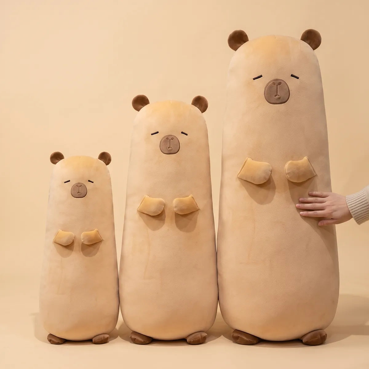 The three published heights of the big capybara stuffed animal plush lined up with an adult hand resting on the tallest one.