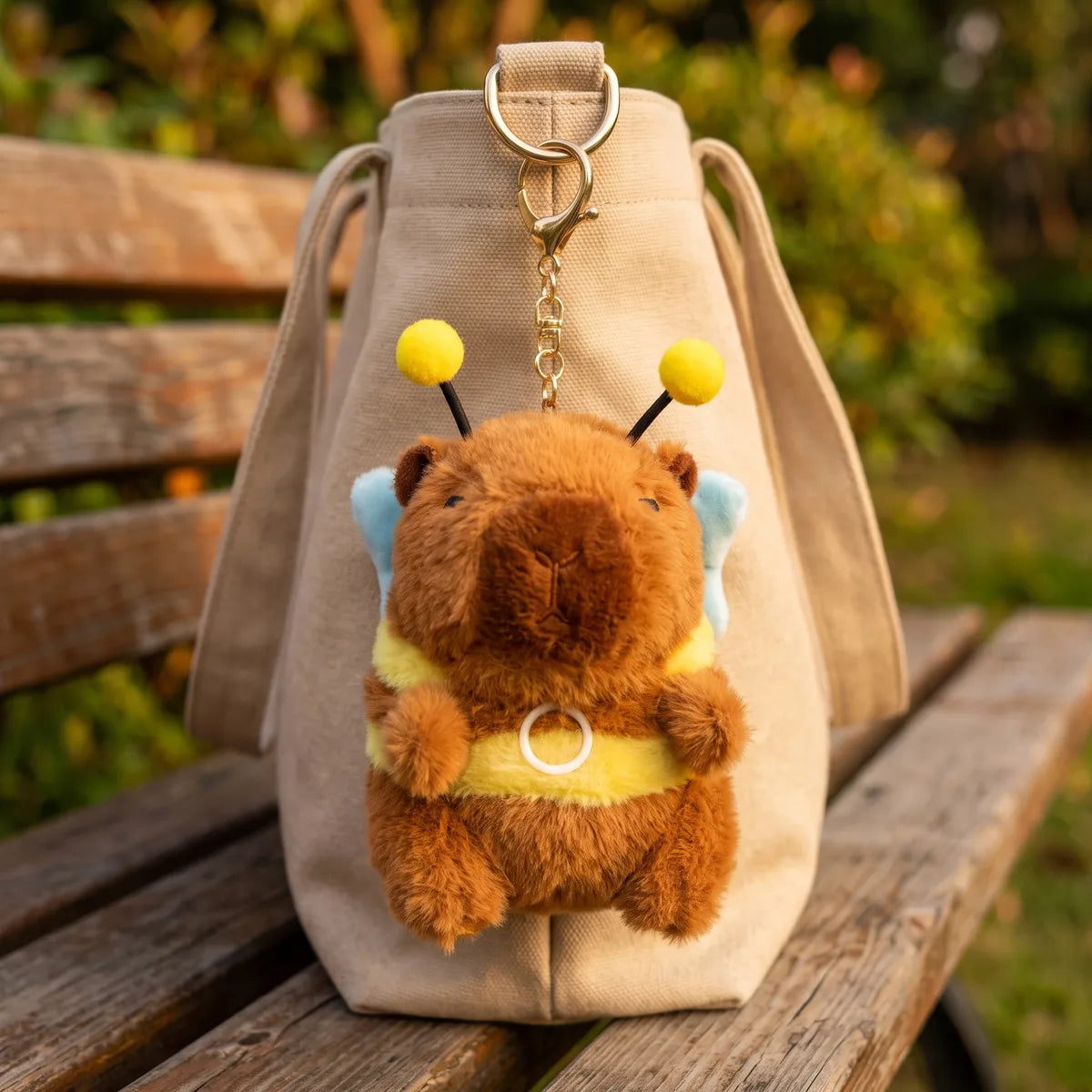 Capybara bee keychain plush hanging front on by its gold clasp from the metal D ring of a canvas tote, the pale blue wings showing on either side