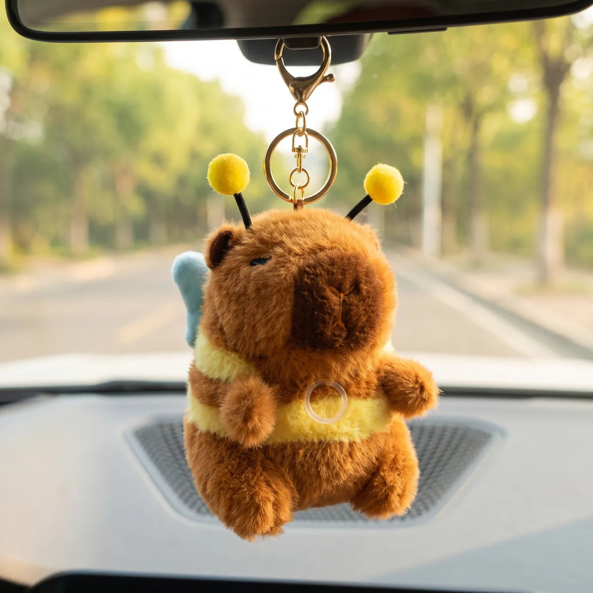 Capybara bee keychain plush hanging from a car rear view mirror with a tree lined road in the background