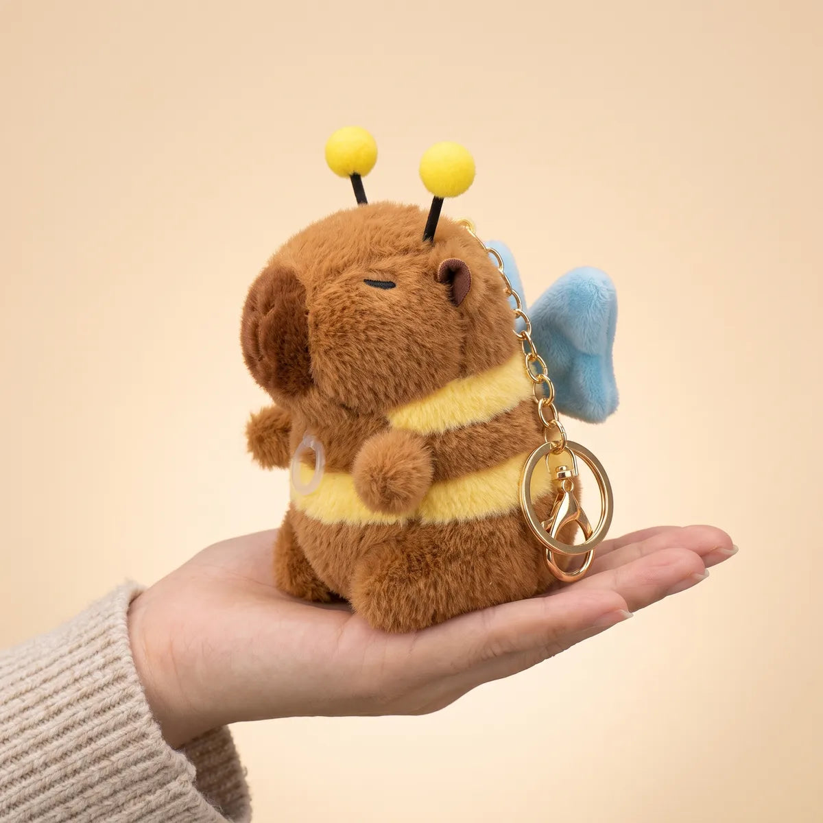 Capybara bee keychain plush sitting upright on an open palm with its gold clasp hanging over the side