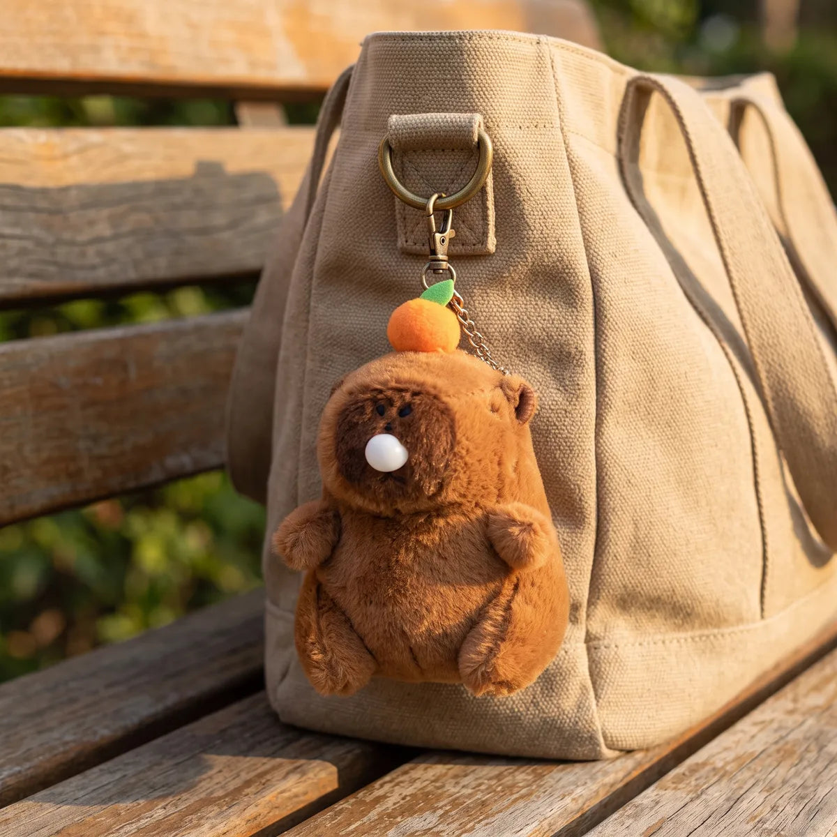 Capybara blowing bubble keychain plush clipped to the D ring of a beige canvas tote bag on a wooden bench.