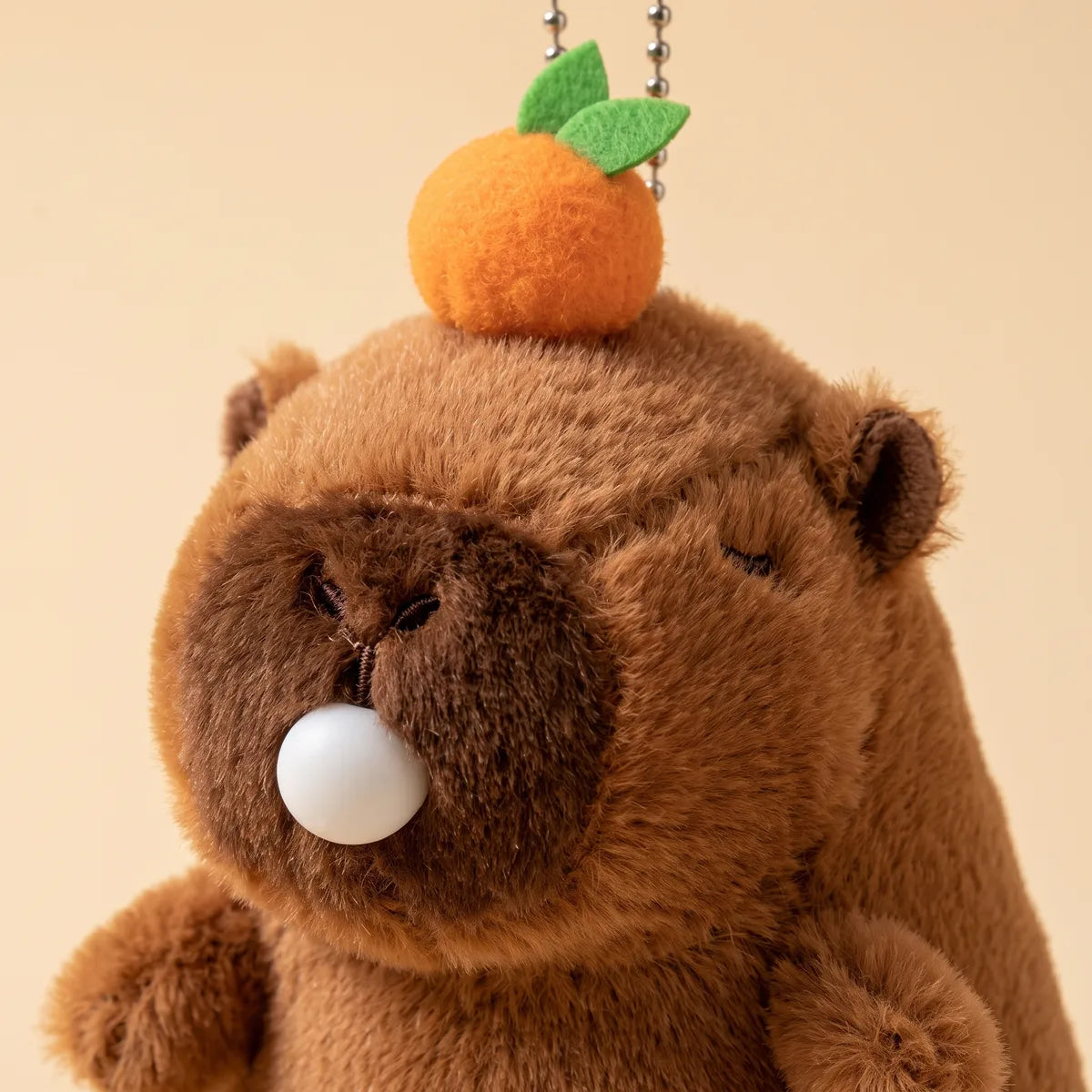 Close up of the capybara blowing bubble keychain plush head, the orange pom pom with felt leaves threaded on the ball chain above its embroidered eyes