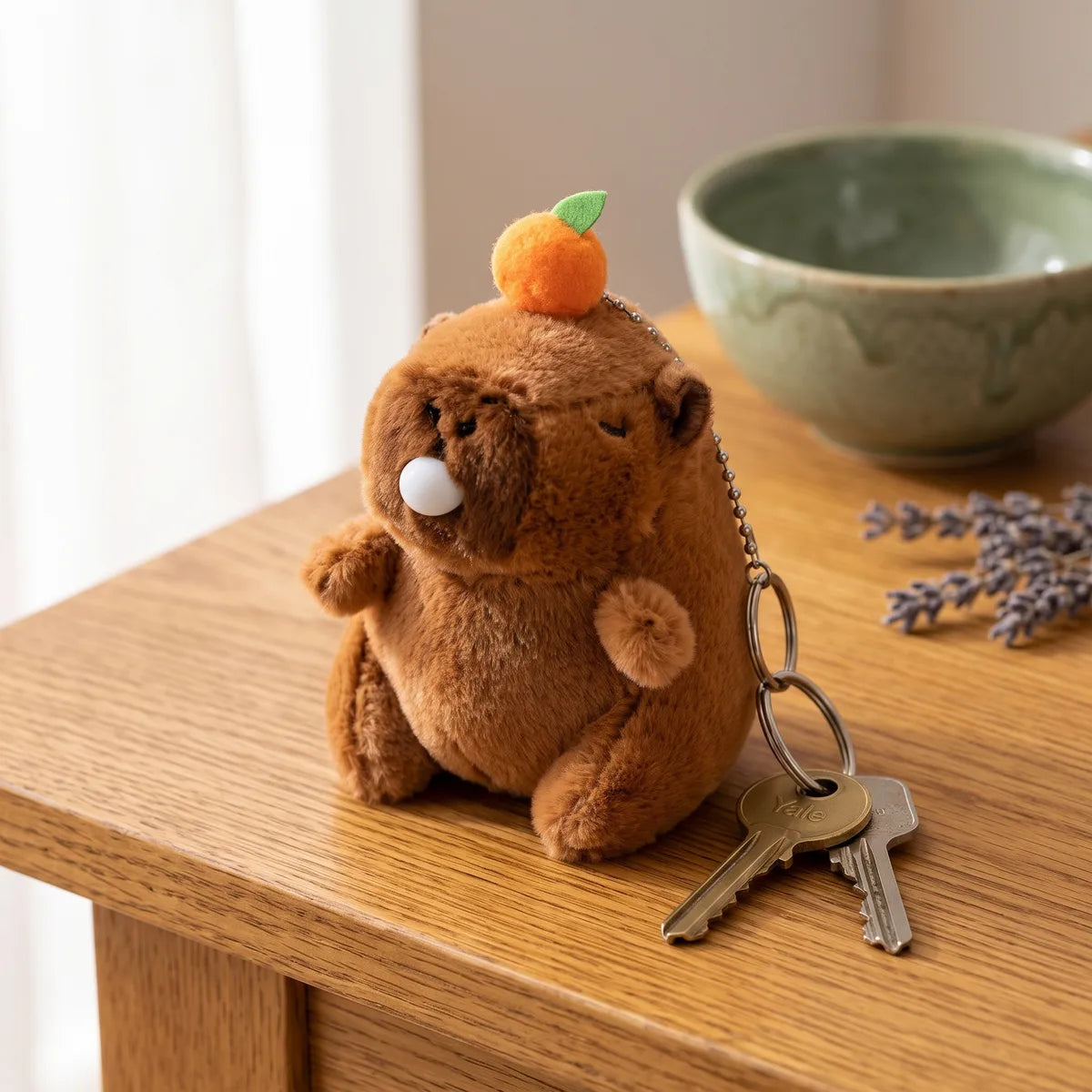 Capybara blowing bubble keychain plush resting on a wood table beside a brass key and a silver key on its split ring