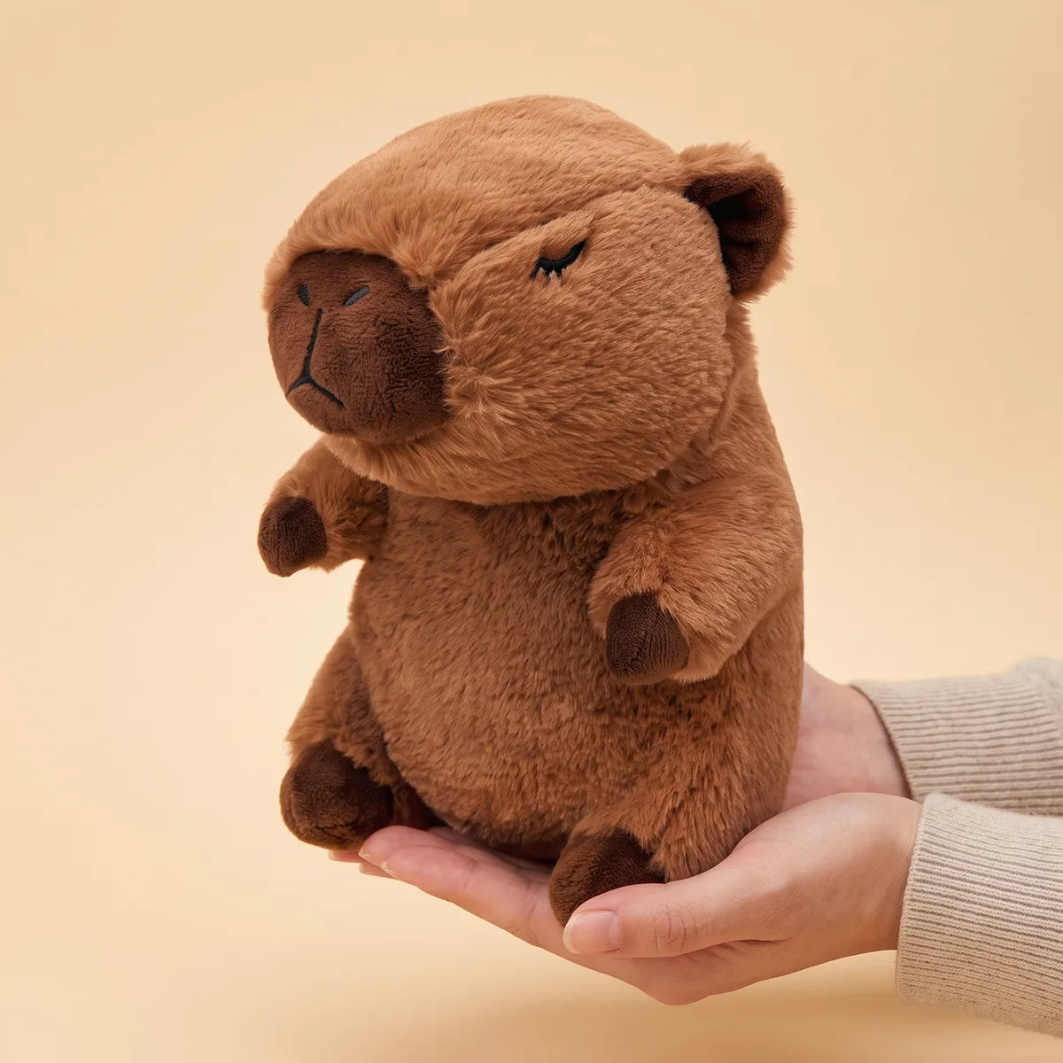 Capybara breathing plush resting across two cupped hands, showing its 13 inch seated height against an adult palm.