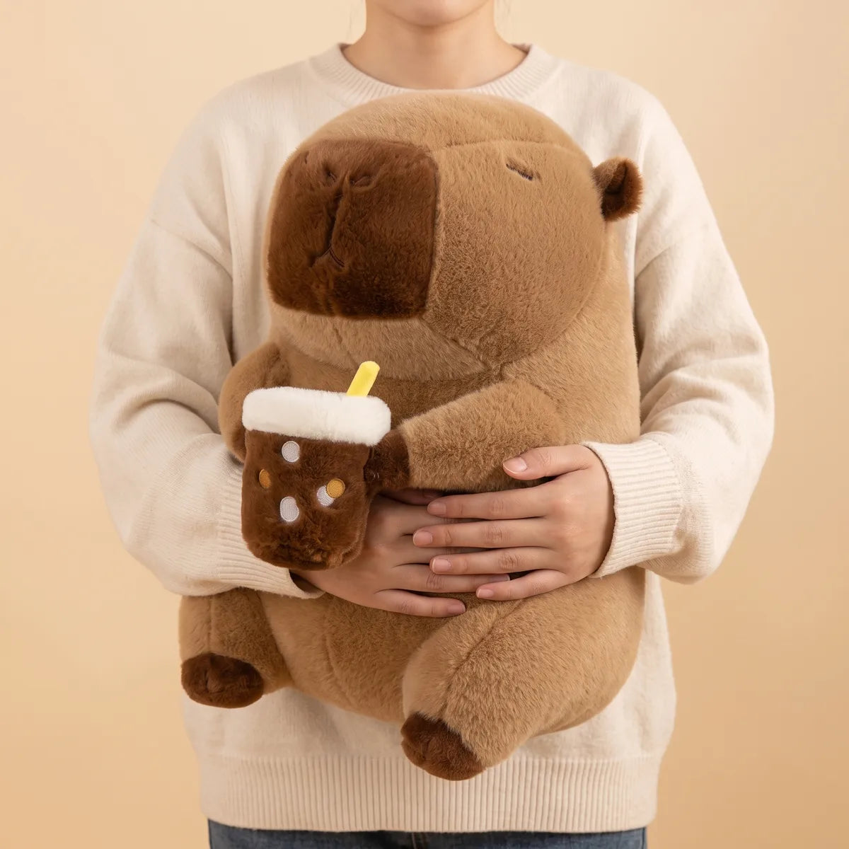 Capybara drinking boba plush held at waist height against a torso, both hands under its belly and the bubble tea cup still in its paws