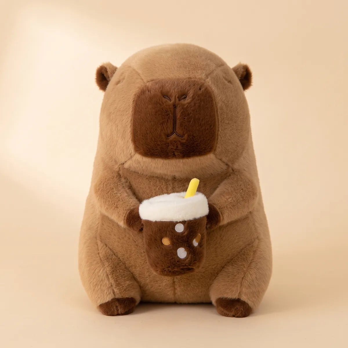 Capybara drinking boba plush seated front on, holding its foam topped bubble tea cup with a yellow straw.