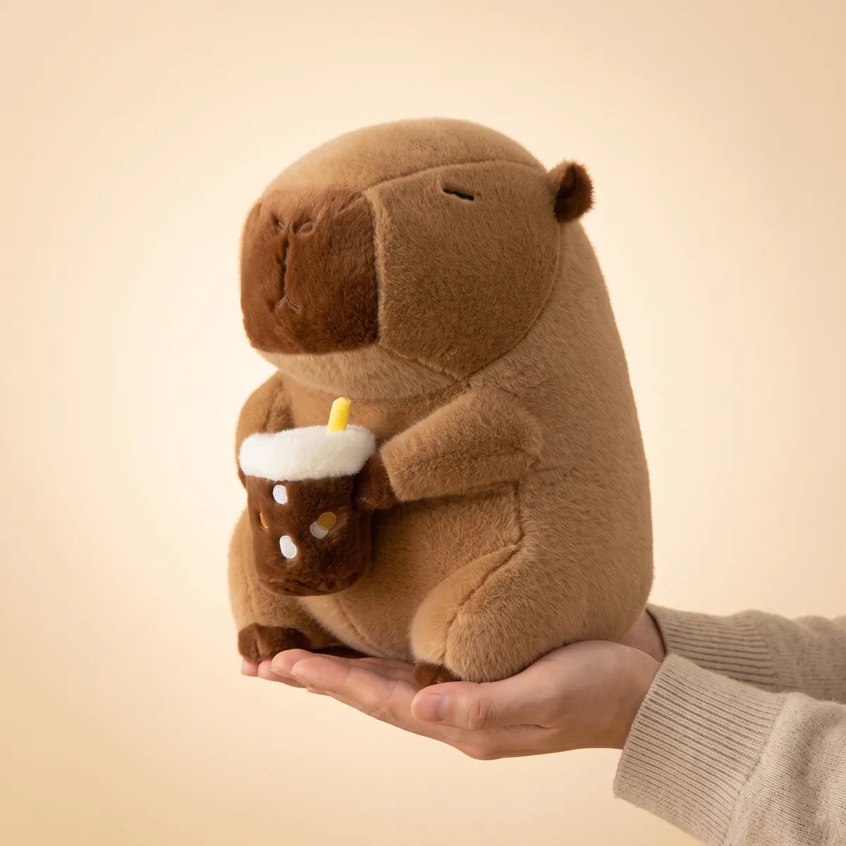 Capybara drinking boba plush resting on one open hand, seen from the side with the boba cup in both paws.