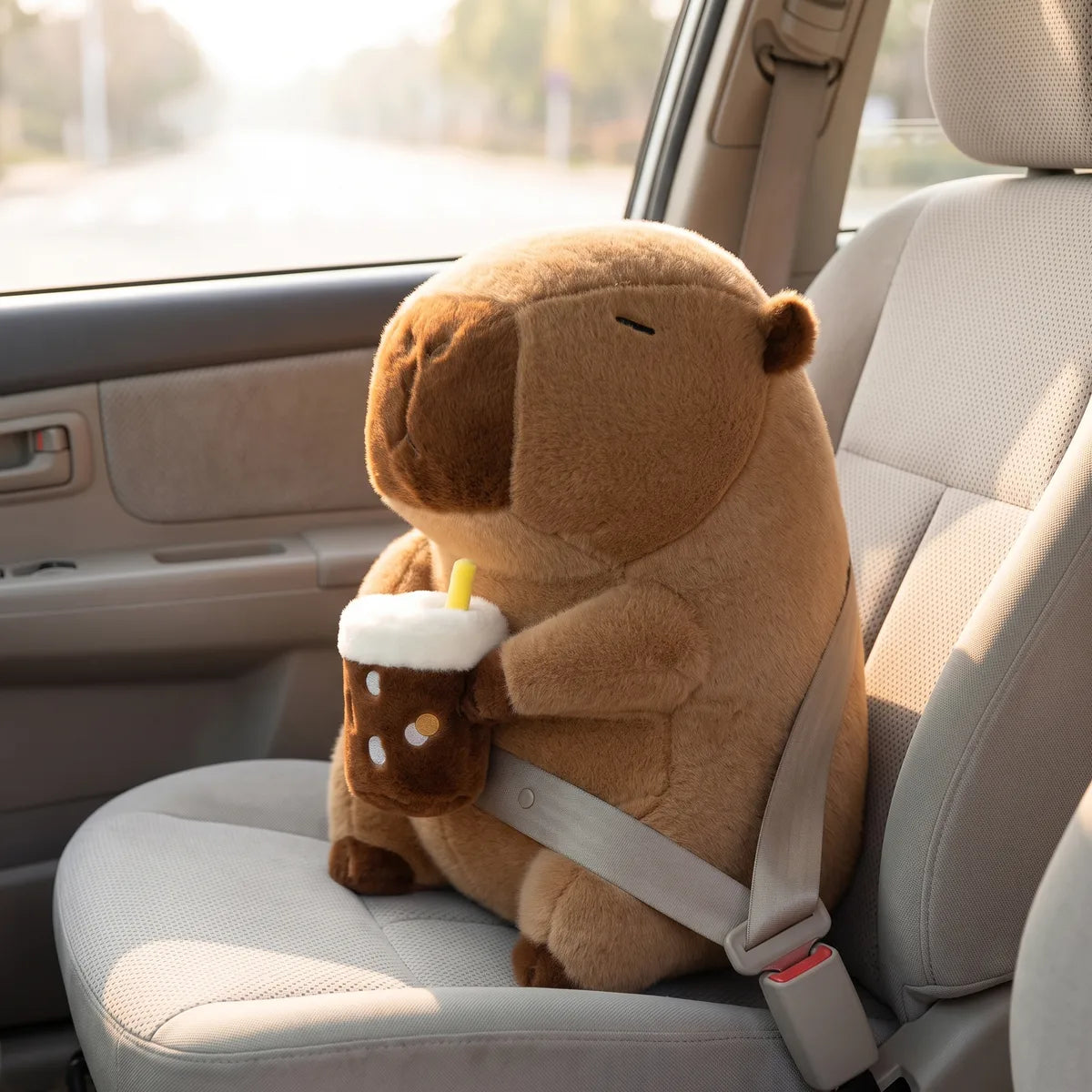 Capybara drinking boba plush strapped into a car passenger seat, the seatbelt running diagonally across its body
