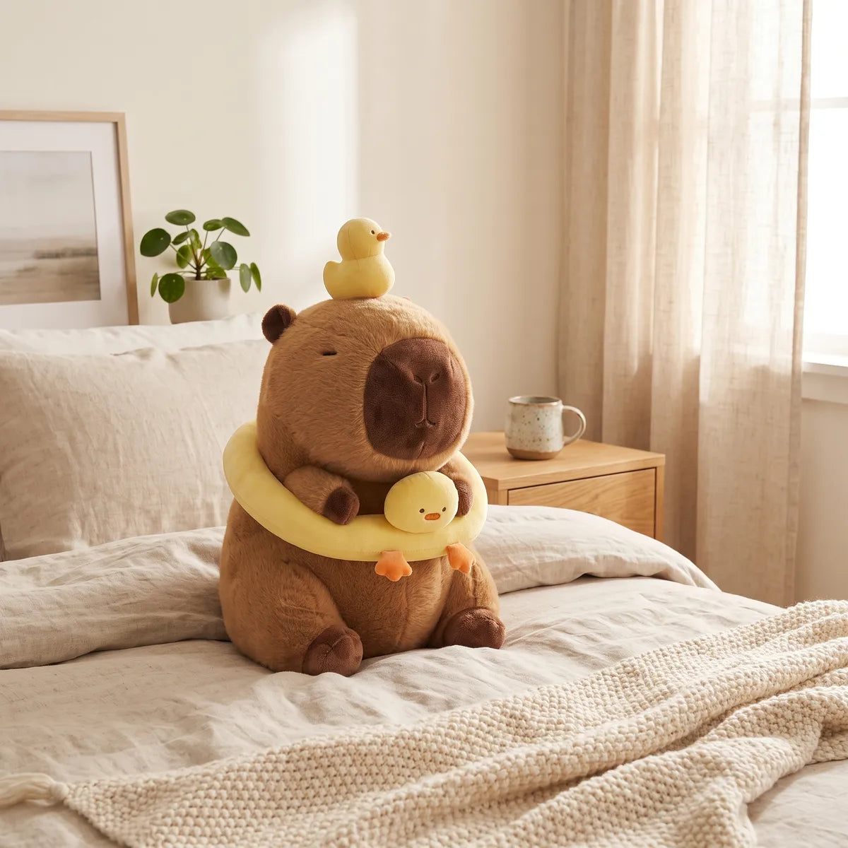 Capybara duck plush seated on a made bed with its yellow buoy in place, a leafy plant behind and a mug at the side