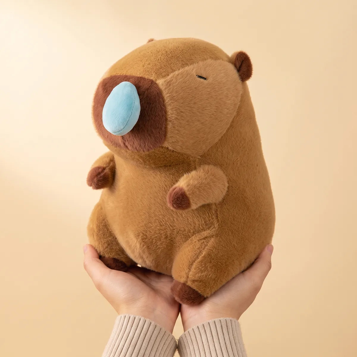 Two hands holding the capybara snot plush to show its real size against adult palms