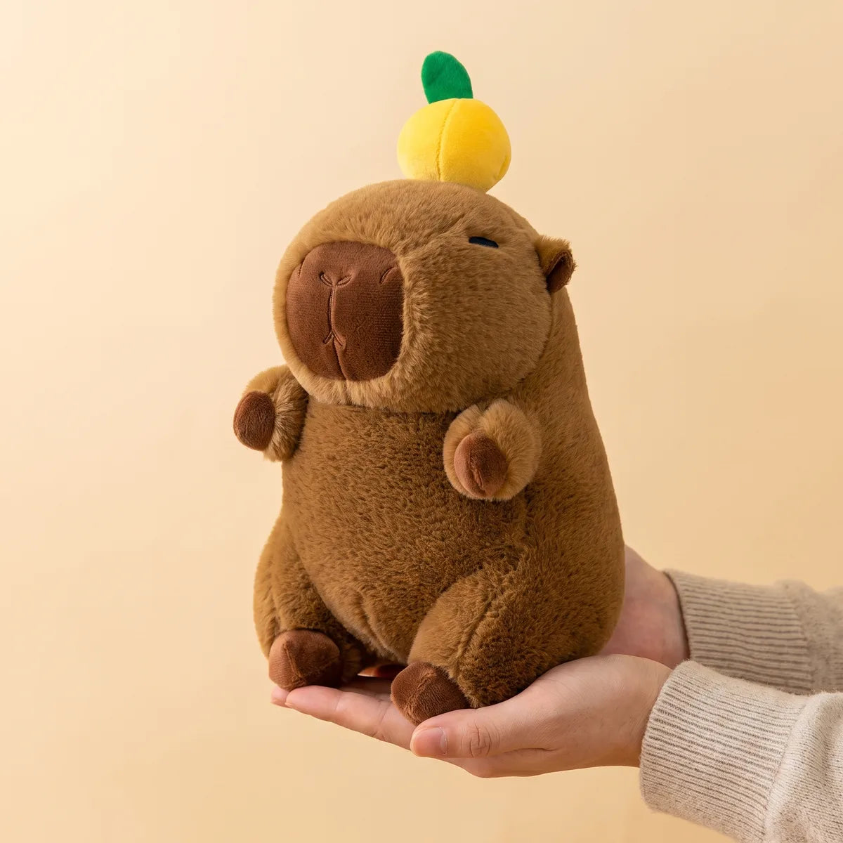 Hands cradling the 9 in capybara with orange on head plush to show its real size