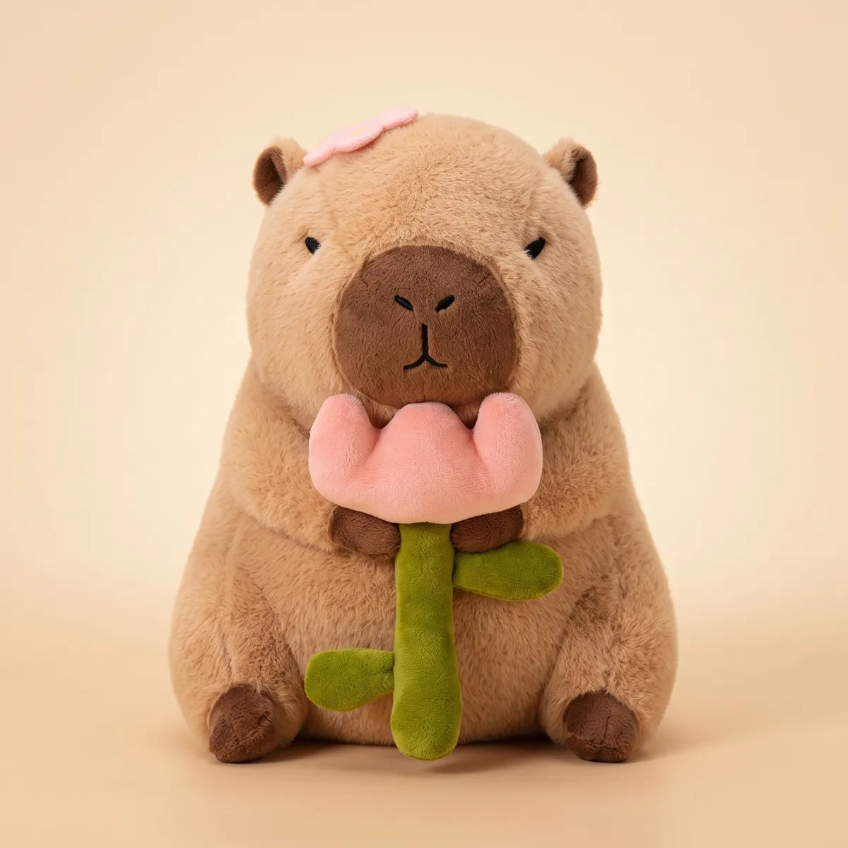Front view of the sandy brown capybara with rose plush holding a fat rose pink bloom in both paws