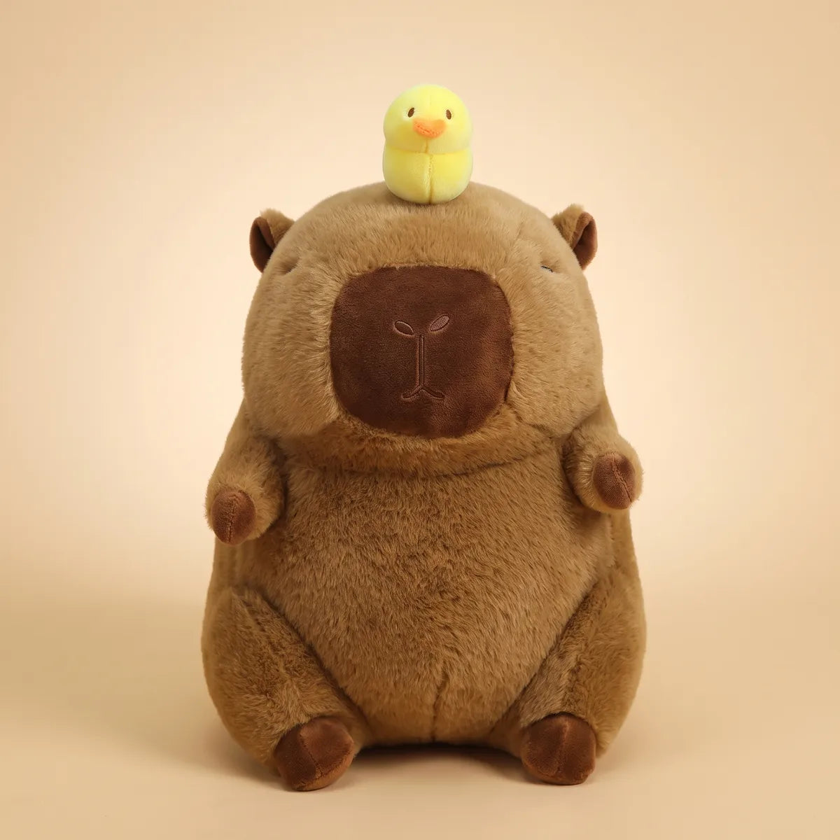 Front view of the brown capybara with duck on head plush sitting upright on a cream studio backdrop