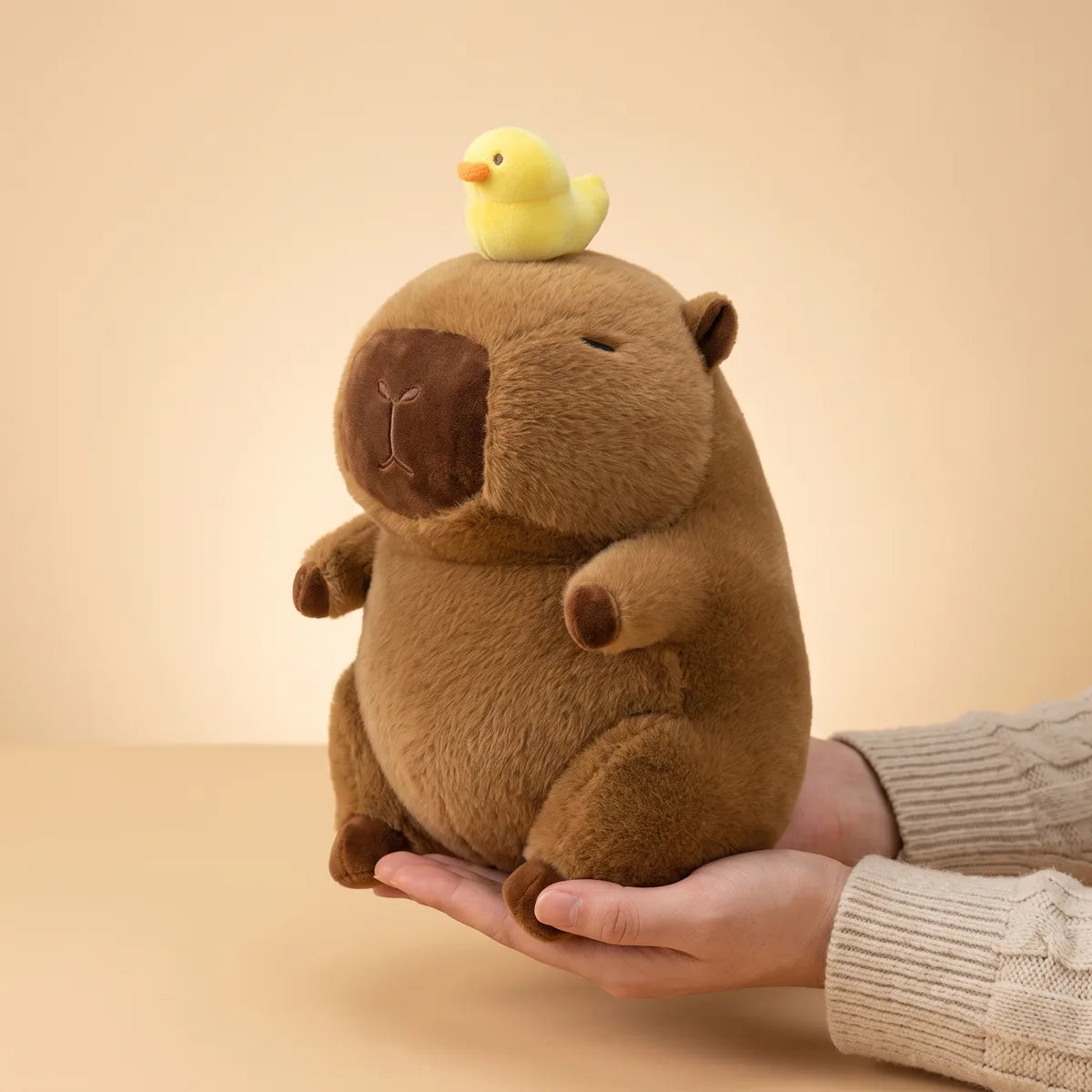 Capybara with duck on head plush resting on an open hand, its brown back feet on the palm and its body rising well above the fingers