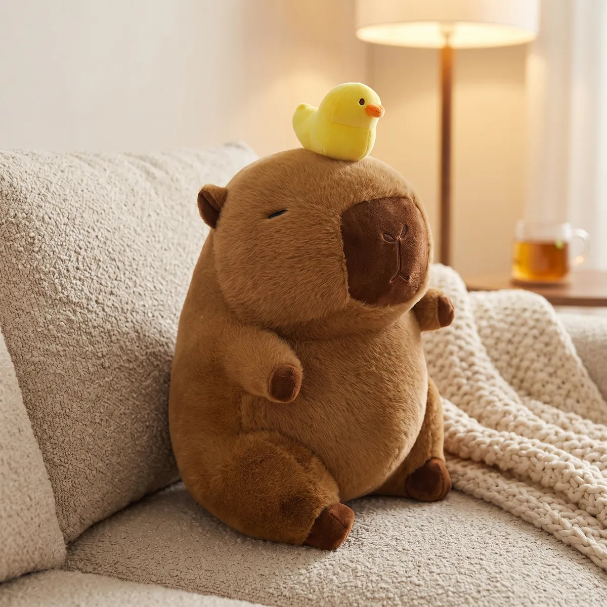 Capybara with duck on head plush resting on a boucle sofa next to a warm floor lamp and a glass of tea