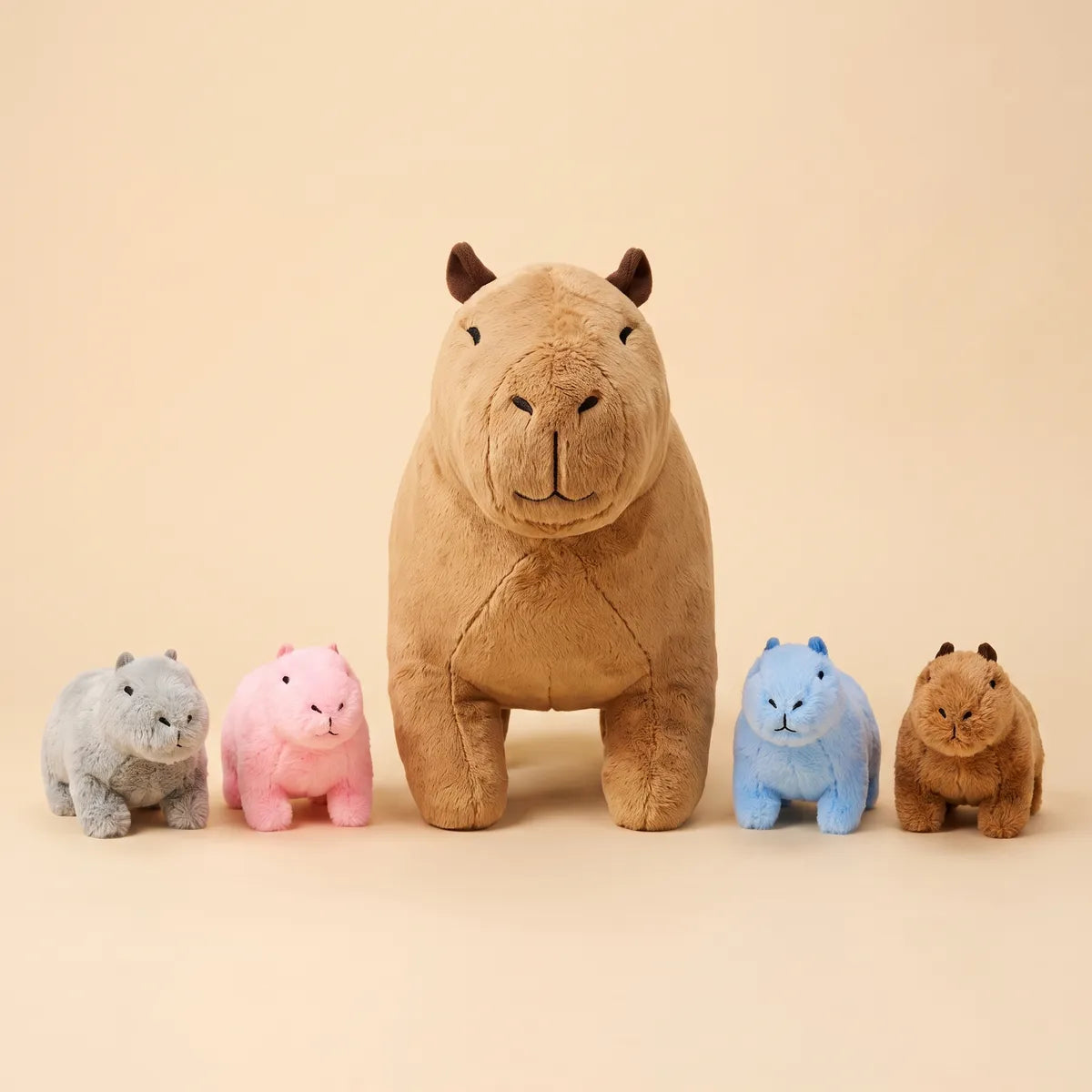 All five pieces of the mother capybara plush set lined up on cream: grey, pink, blue and brown babies beside the tan mother