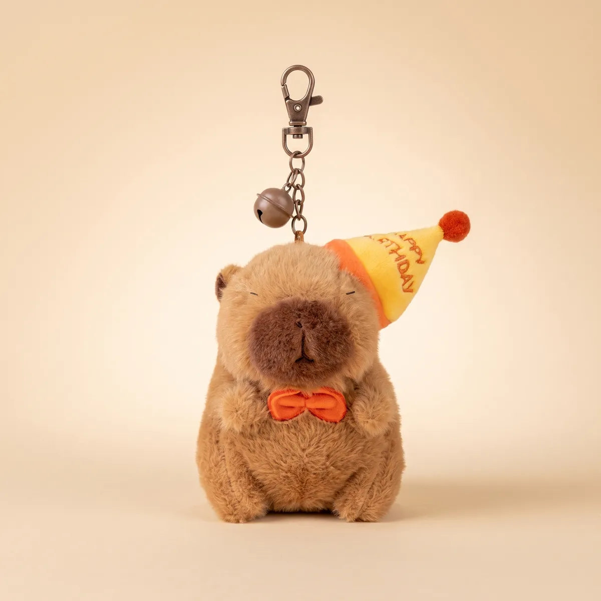 Front view of the birthday capybara keychain hanging from its chain, bell and lobster clasp on a beige backdrop