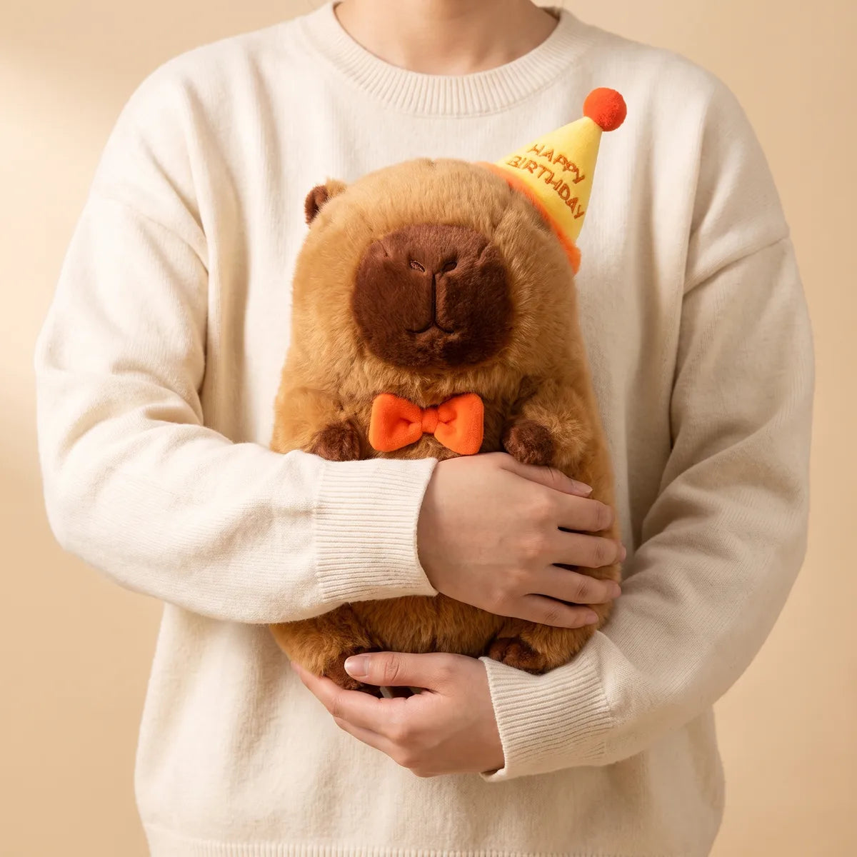 Person in a cream sweater carrying the 13 in happy birthday capybara plush in both arms