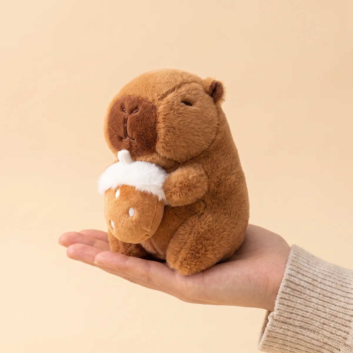 Hazelnut mini capybara plush sitting inside an open palm with its nut resting on the fingers