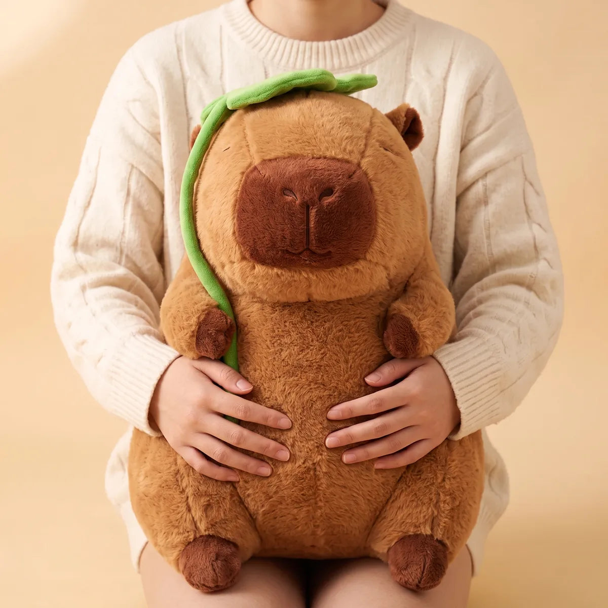 Adult in a cable knit sweater holding the jumbo capybara plush upright on their lap with both hands.