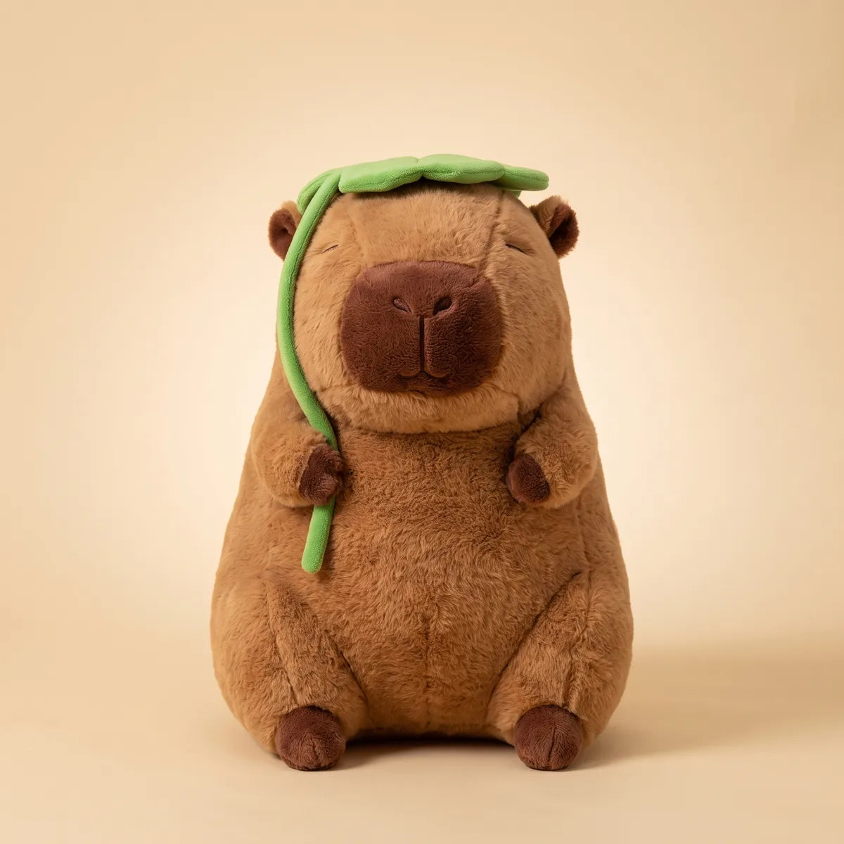 Jumbo capybara plush seated front on against a cream studio backdrop with the green leaf over its head.
