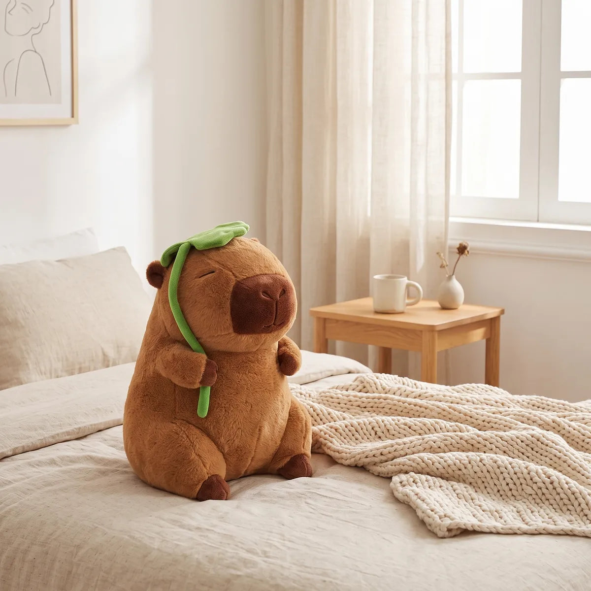 Jumbo capybara plush sitting upright on a duvet with its sewn on green leaf over its head and a chunky knit throw spread beside it
